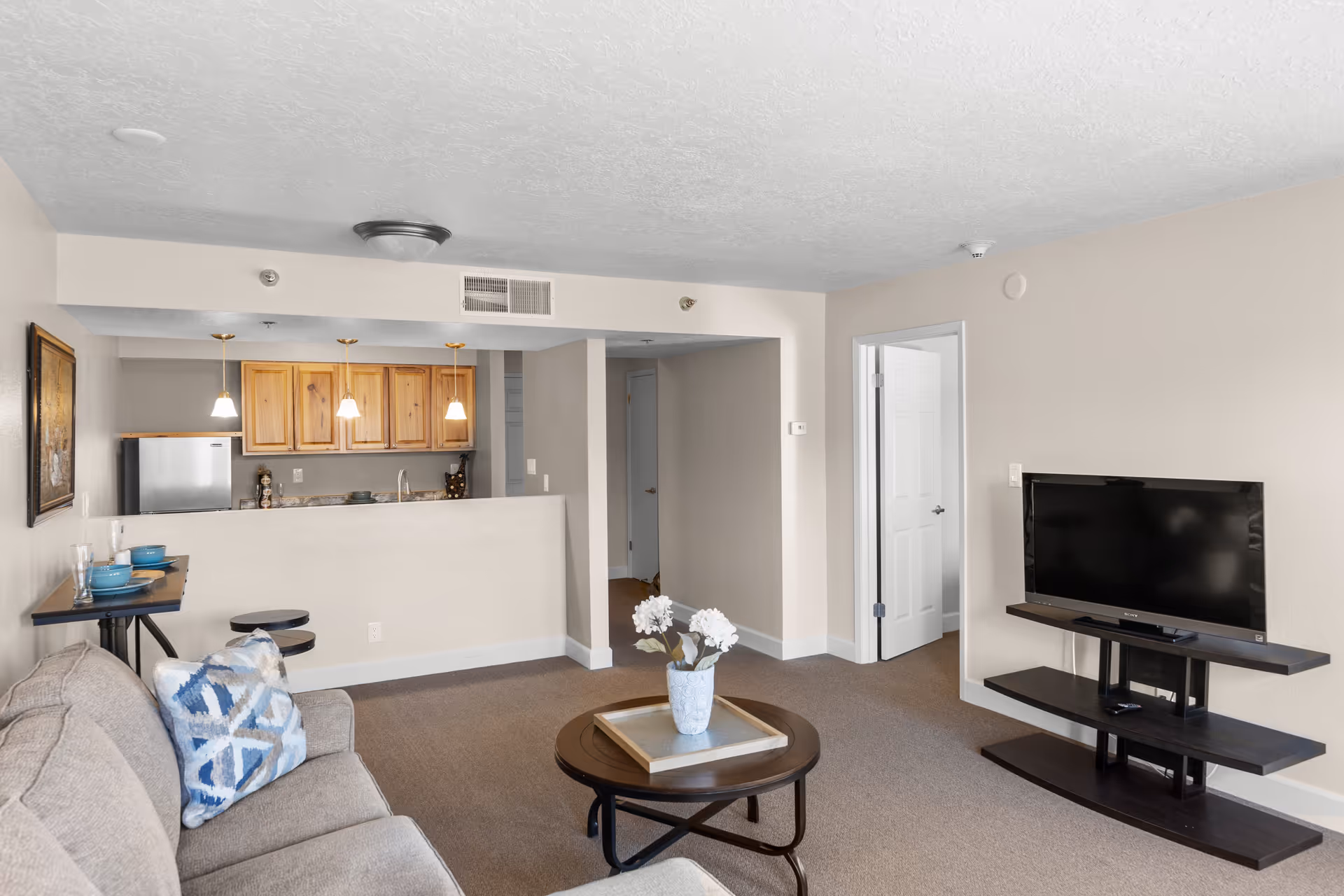 A living room area with a beige sofa adorned with a blue and white patterned pillow, a round wooden coffee table with a vase of white flowers, and a flat-screen TV on a dark wooden stand. In the background, there is an open kitchen with wooden cabinets, a refrigerator, and three pendant lights hanging above a counter with two stools. The walls are painted light beige and the carpet is brown.