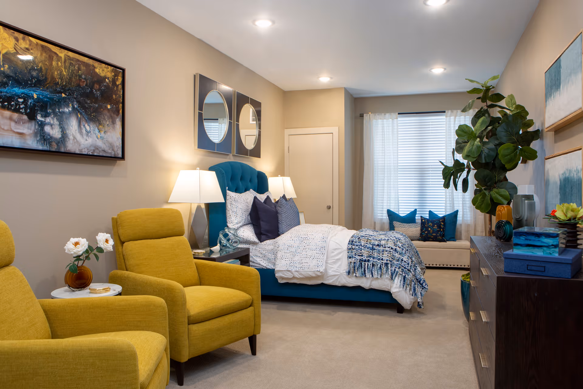 A cozy bedroom featuring a teal upholstered bed with white and blue bedding, two yellow armchairs, a dark wooden dresser with decorative items, a large green plant, and wall art. The room is softly lit with two bedside lamps and natural light coming through a window with white curtains.