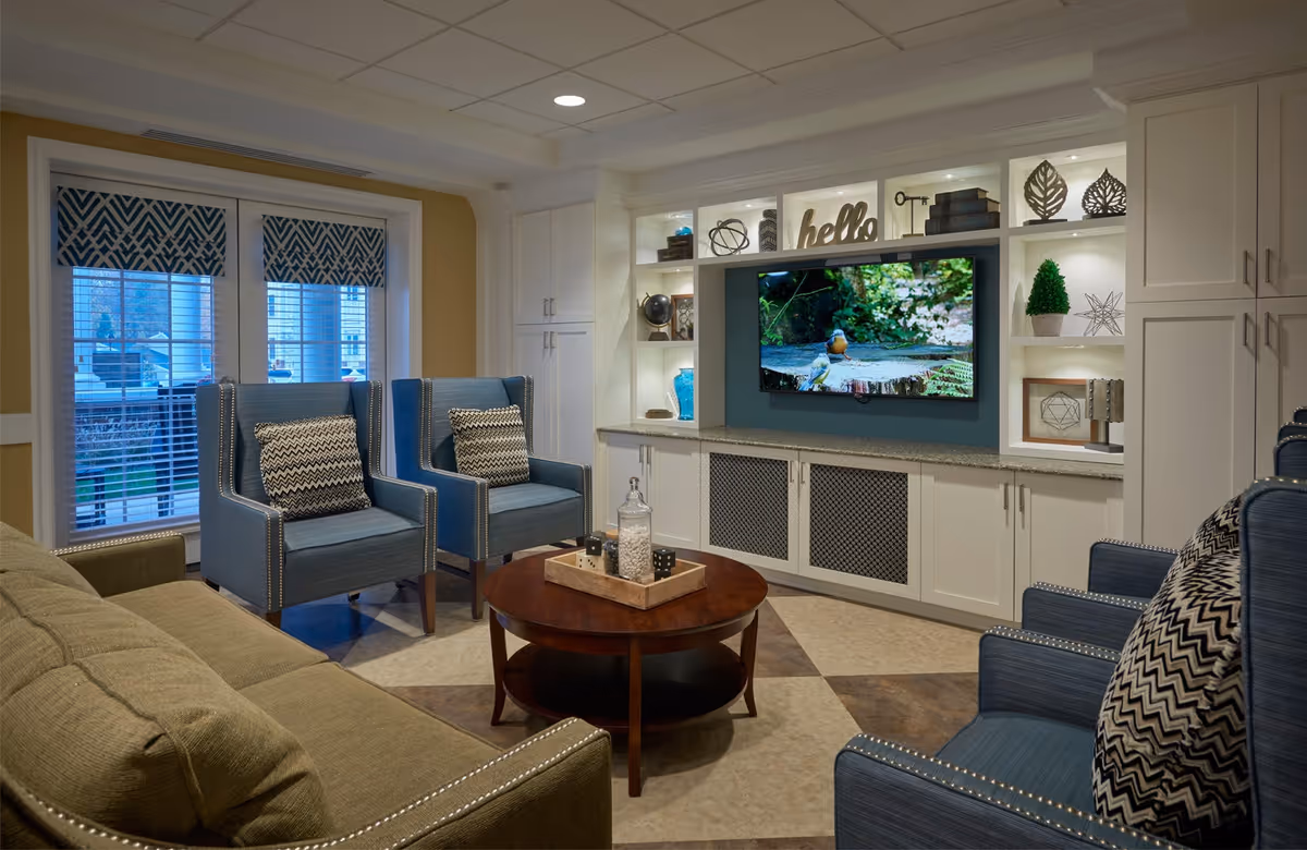 A cozy living room area with a beige sofa and four blue armchairs arranged around a round wooden coffee table. The back wall features built-in white cabinetry with shelves displaying decorative items and a mounted flat-screen TV showing a nature scene. Large windows with patterned valances allow natural light into the room.