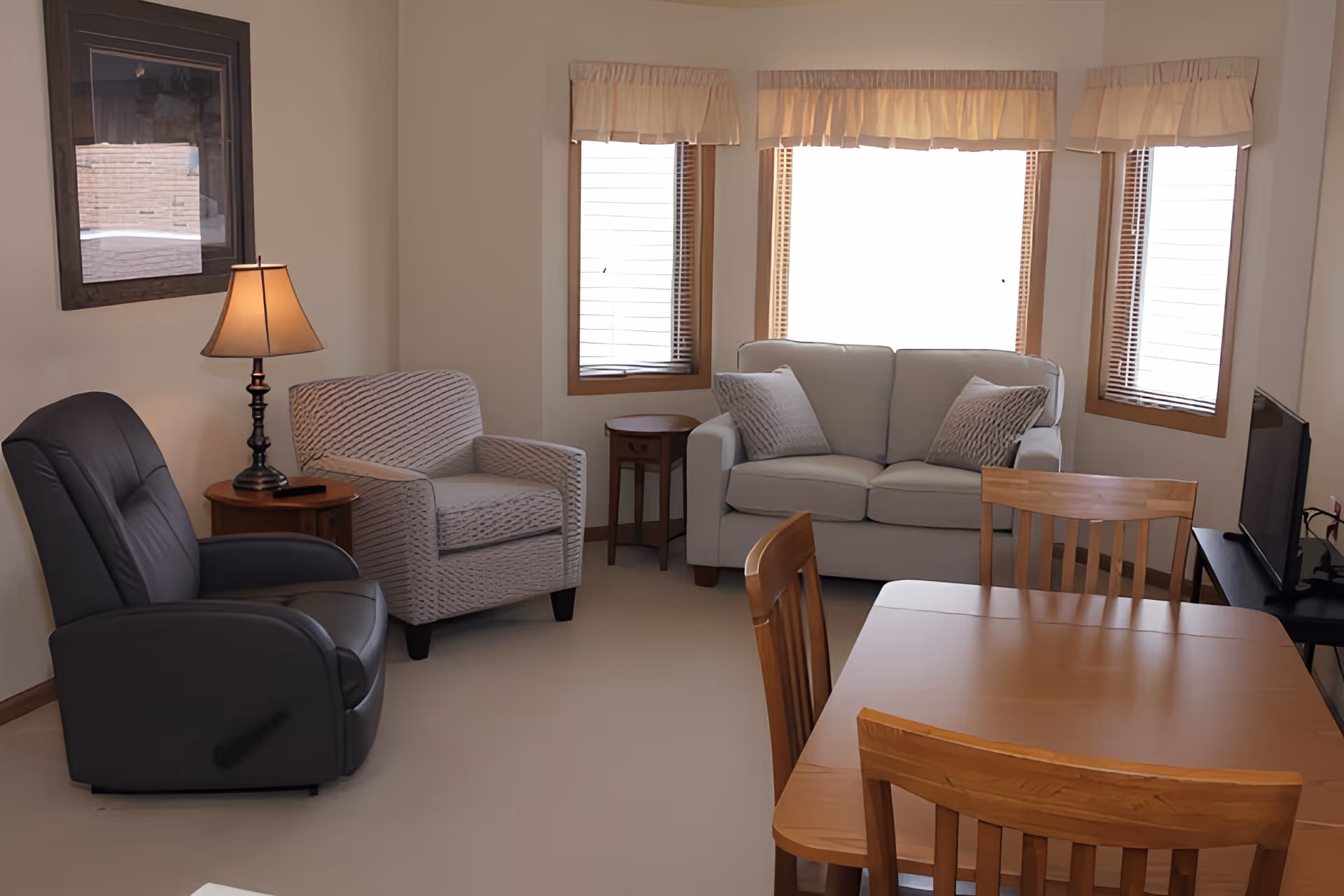 A cozy living room area with a gray sofa, two armchairs, a wooden side table with a lamp, a wooden dining table with chairs, and a flat-screen TV on a stand. The room has three windows with beige valances and wooden blinds.