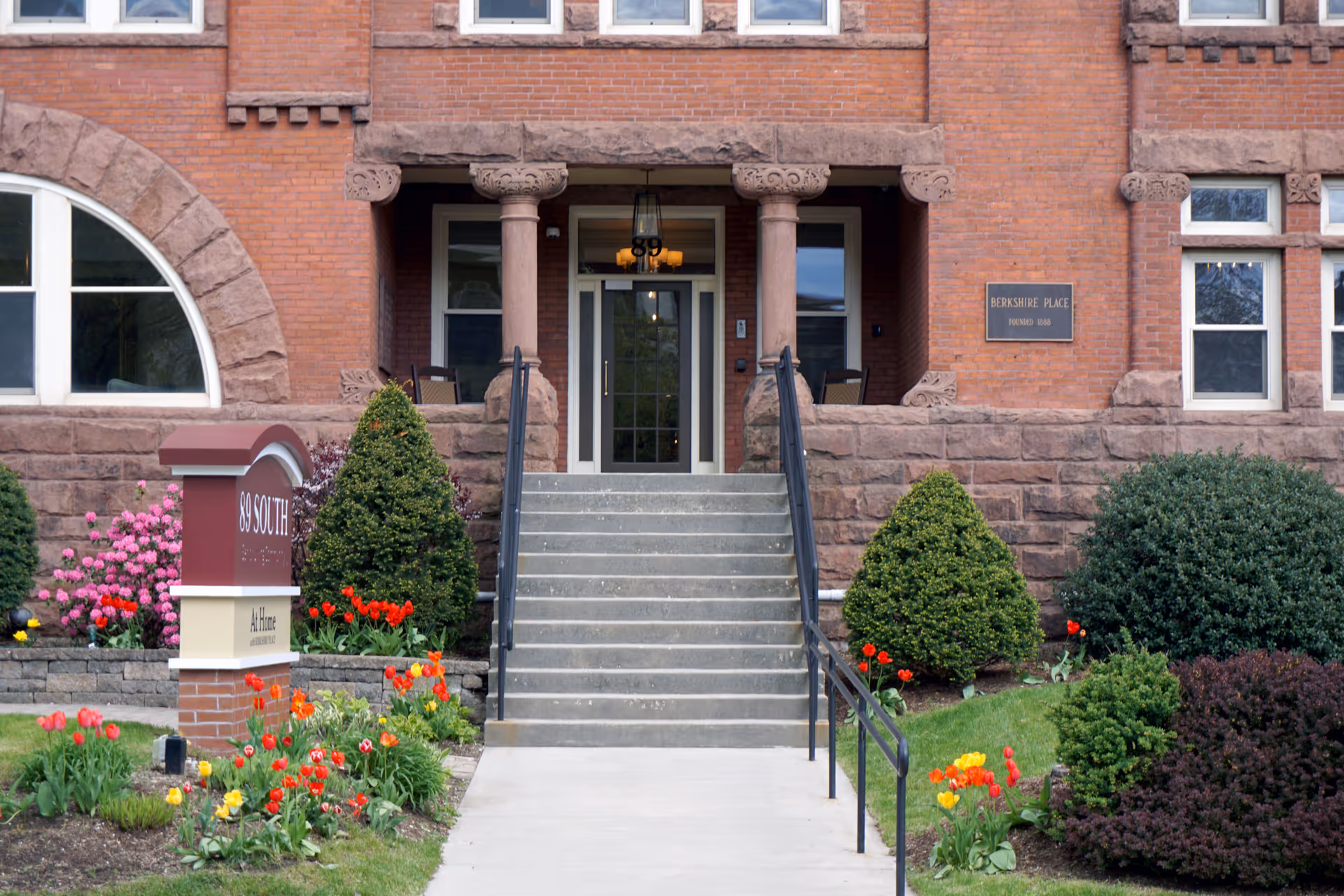 Front entrance of a brick building with stone columns and steps leading up to a glass door. There are well-maintained bushes and colorful flowers on either side of the walkway. A sign in front reads '89 South' and 'Berkshire Place'.