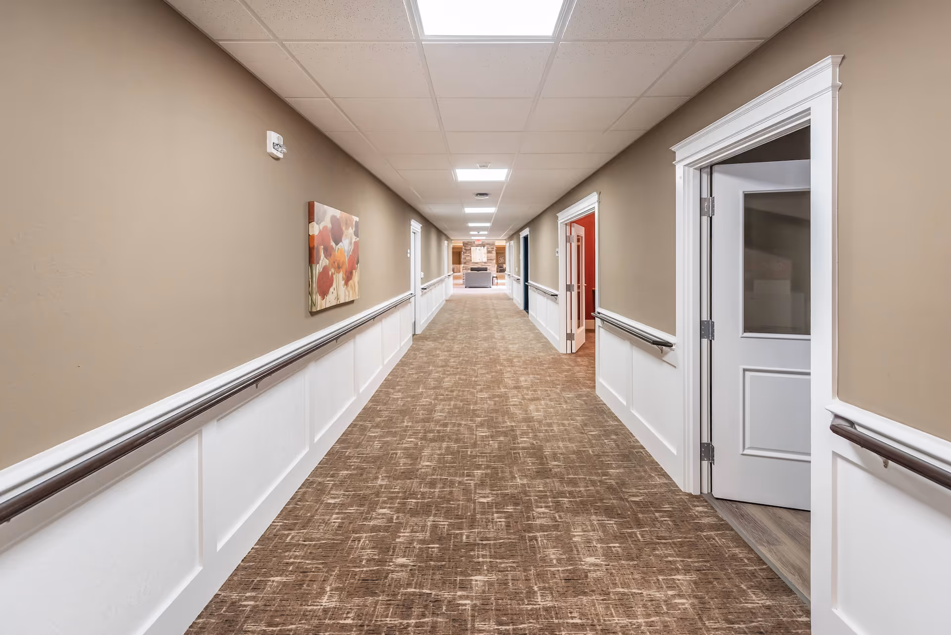 Long hallway in a senior living facility with beige walls, white wainscoting, brown patterned carpet, handrails on both sides, and several doors along the hallway. A colorful floral painting is hung on the left wall, and the hallway leads to a seating area with a fireplace in the distance.