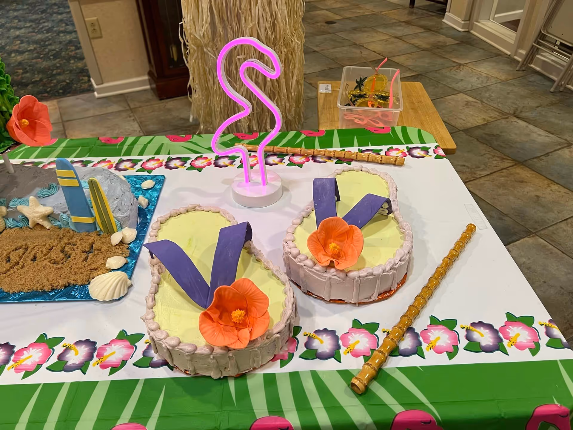 Tropical-themed table display with flip-flop-shaped cakes, a neon flamingo light, and beach decorations on a floral tablecloth inside the facility.