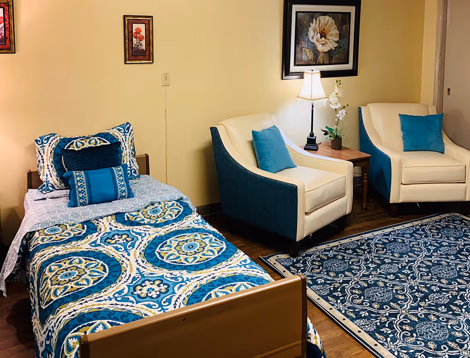 A decorated bedroom with a twin bed dressed in blue patterned bedding, two cream-and-teal armchairs, a side table with lamp, and a blue area rug.
