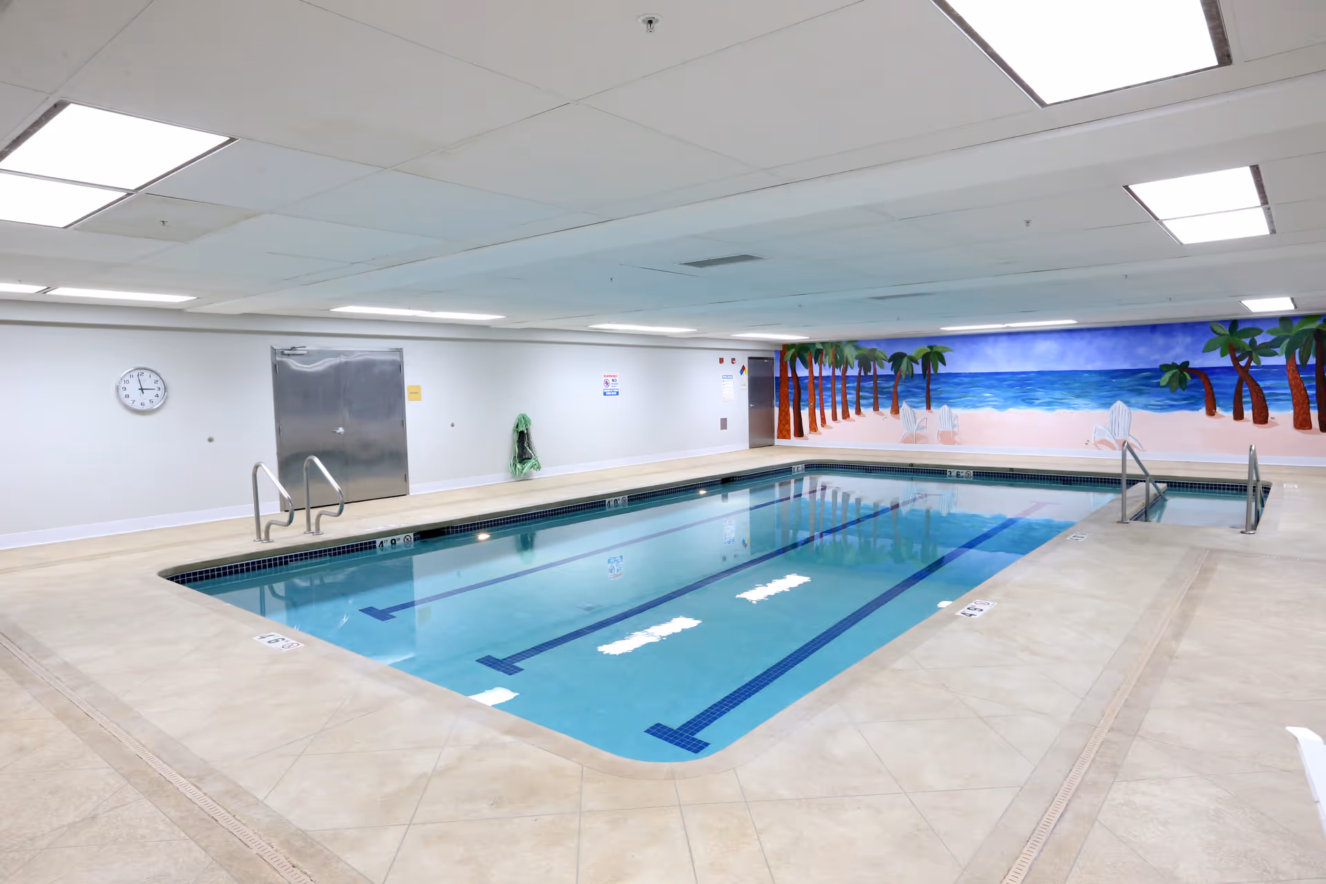 Indoor community swimming pool with lane markings and a beach mural on the far wall.