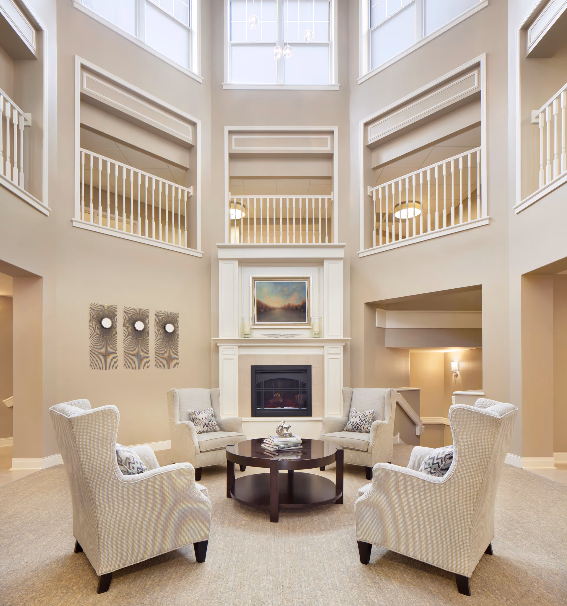 A spacious and elegant living room area with four beige upholstered armchairs arranged around a dark wooden round coffee table. The room features a tall ceiling with large windows, a white fireplace with a framed painting above it, and decorative wall art on the left side. The walls are painted in a soft beige tone, and there are white railings on the upper level overlooking the room.
