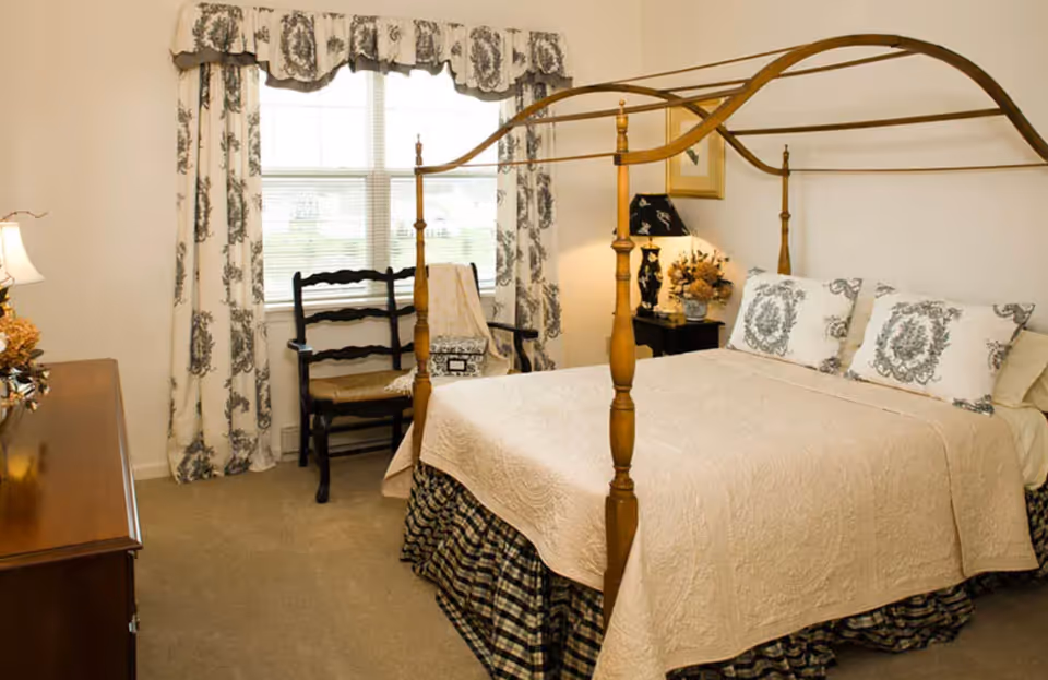 A bedroom with a wooden four-poster bed covered with a beige quilt and matching pillows. There is a window with patterned curtains, a wooden chair with a cushion, a nightstand with a lamp and floral arrangement, and a wooden dresser with a lamp on it.