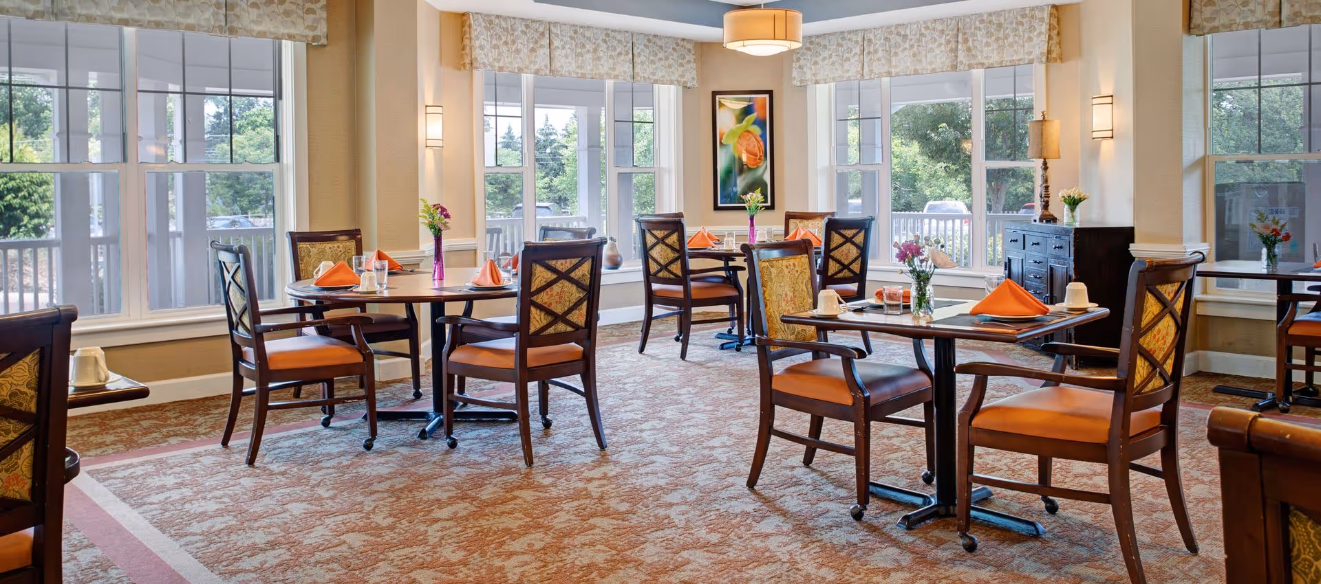 A bright and inviting dining room with several wooden tables and chairs arranged neatly. Each table is set with orange folded napkins, glasses, and small flower vases. Large windows allow natural light to fill the room, and there is a framed colorful artwork on the wall. The room has a warm, patterned carpet and soft lighting from wall sconces and a ceiling fixture.
