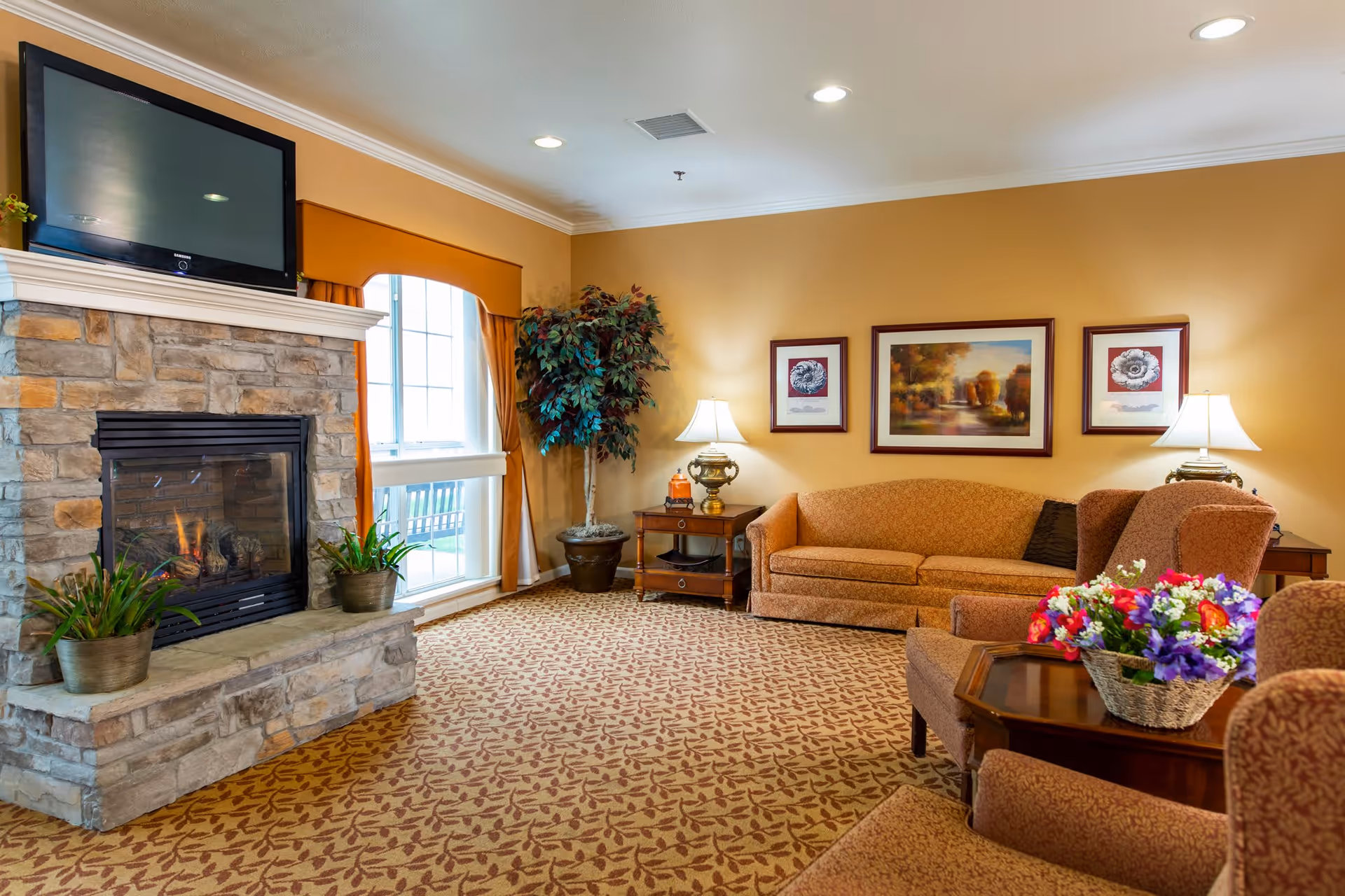 A cozy living room with a stone fireplace, a flat-screen TV mounted above it, a large window with orange curtains, a potted plant, a brown sofa, two armchairs, a wooden coffee table with a basket of colorful flowers, two side tables with lamps, and framed artwork on the wall.