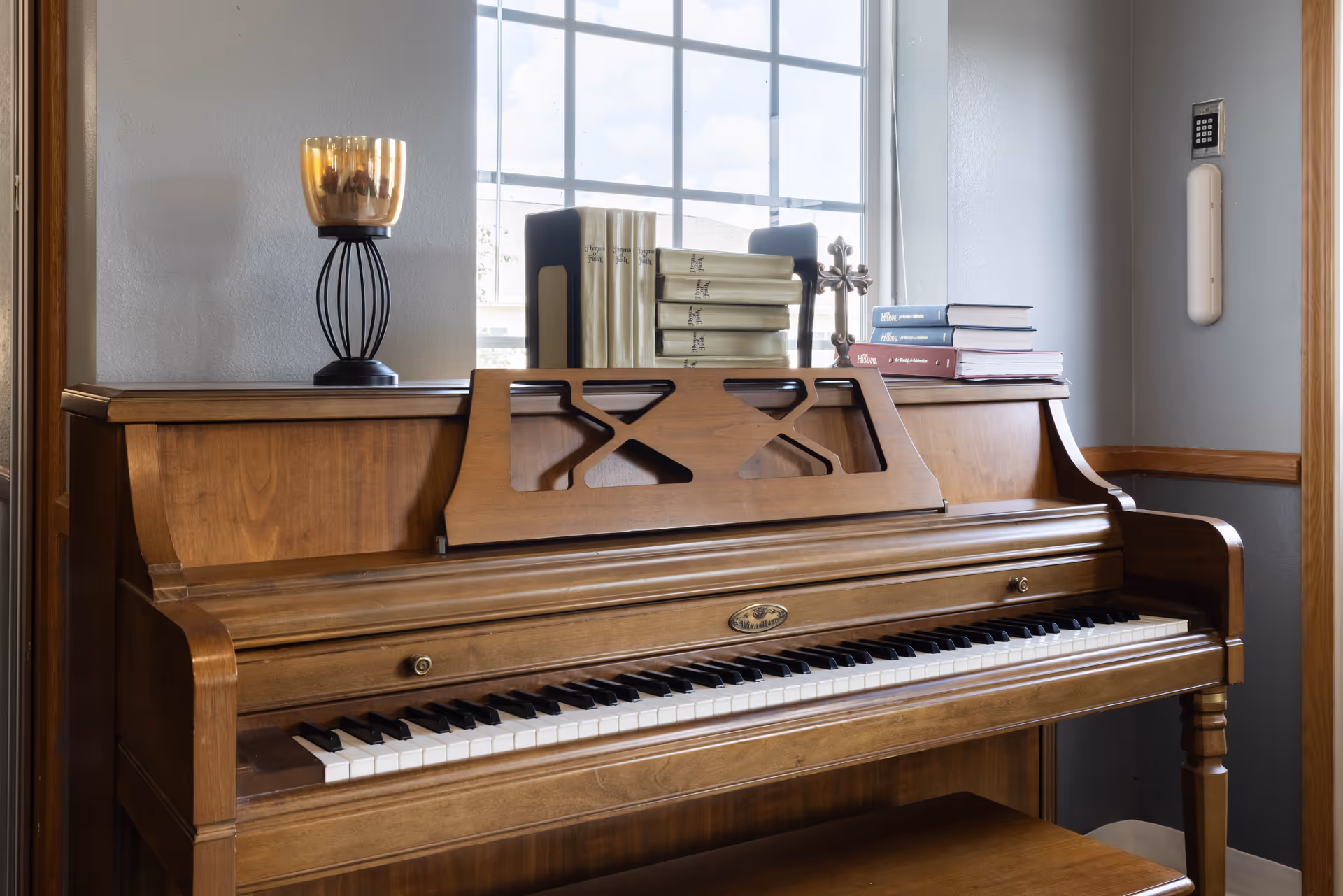 A wooden upright piano with a music stand, books, and a decorative candle holder on top, positioned in front of a window with a grid pattern. The room has light gray walls with wooden trim and a keypad mounted on the wall.