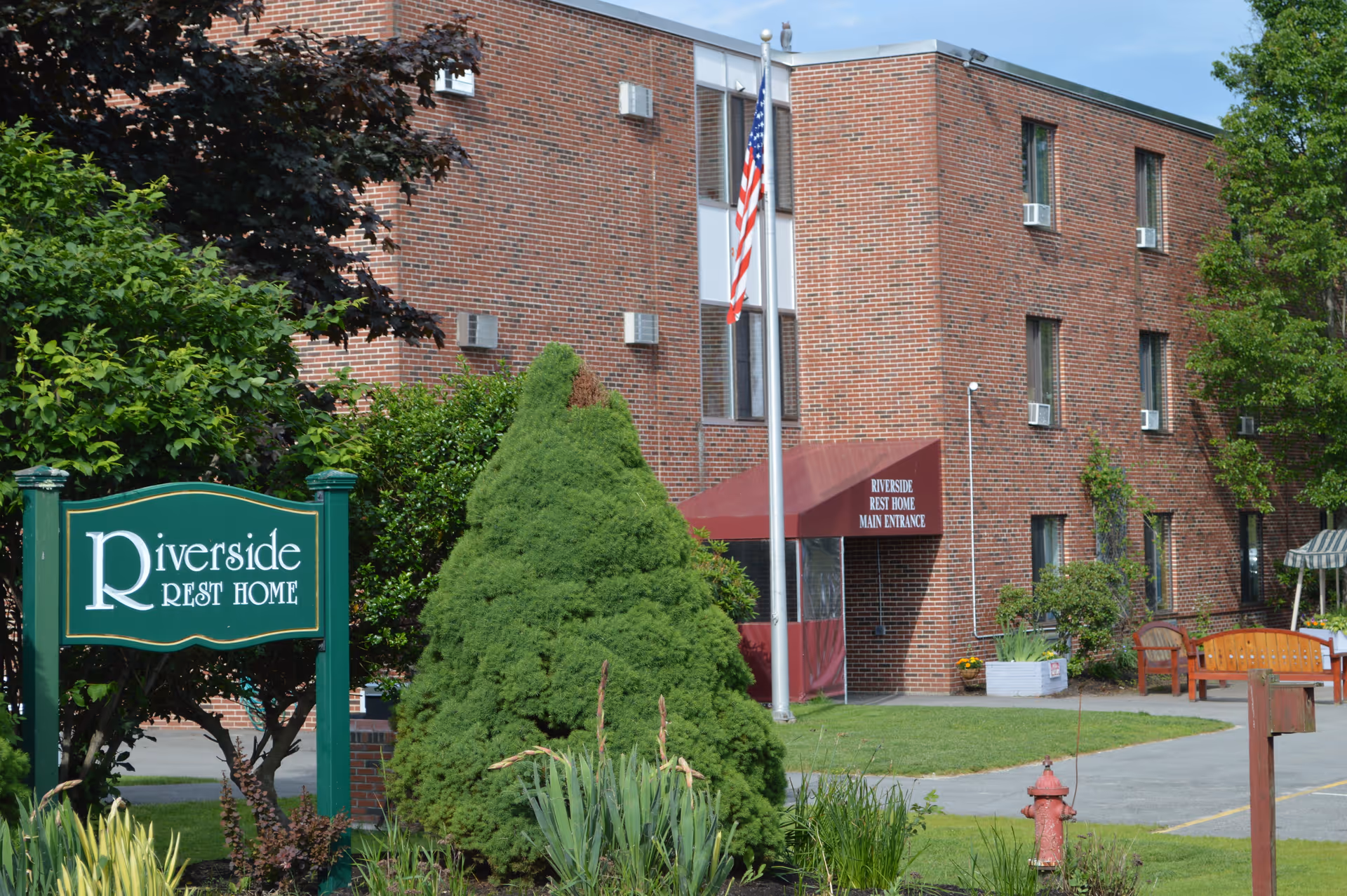 Exterior view of Riverside Rest Home, a three-story brick building with several windows and window air conditioning units. There is a green sign with white text reading 'Riverside Rest Home' near the entrance. A flagpole with an American flag stands in front of the building, which has a maroon awning labeled 'Riverside Rest Home Main Entrance.' The surrounding area includes green bushes, trees, a fire hydrant, and wooden benches on a paved walkway.