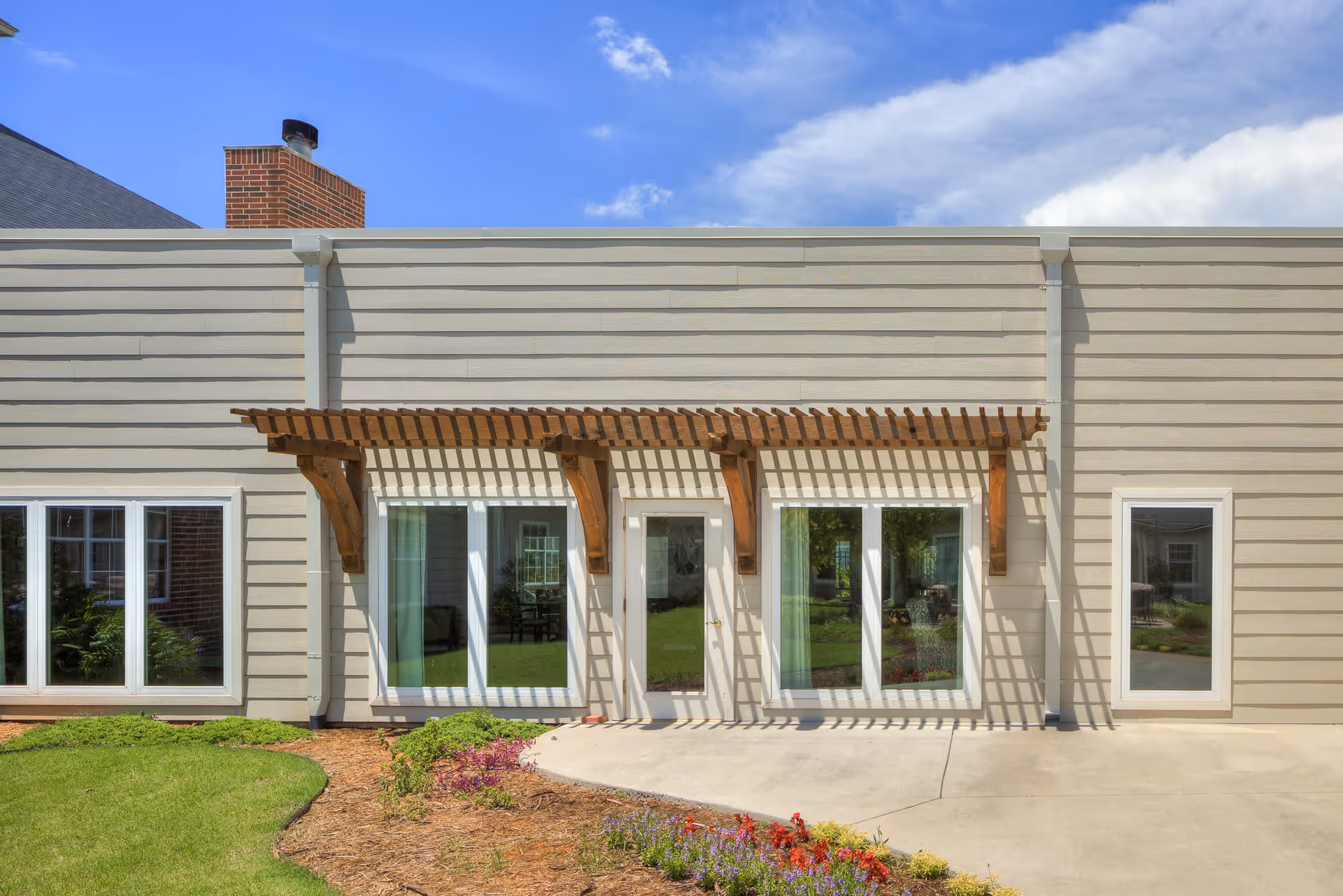 Exterior view of a building with beige siding, featuring a wooden pergola above a central door flanked by large windows. There is a brick chimney on the left side and a landscaped garden with green grass and colorful flowers in front of the building under a blue sky with some clouds.