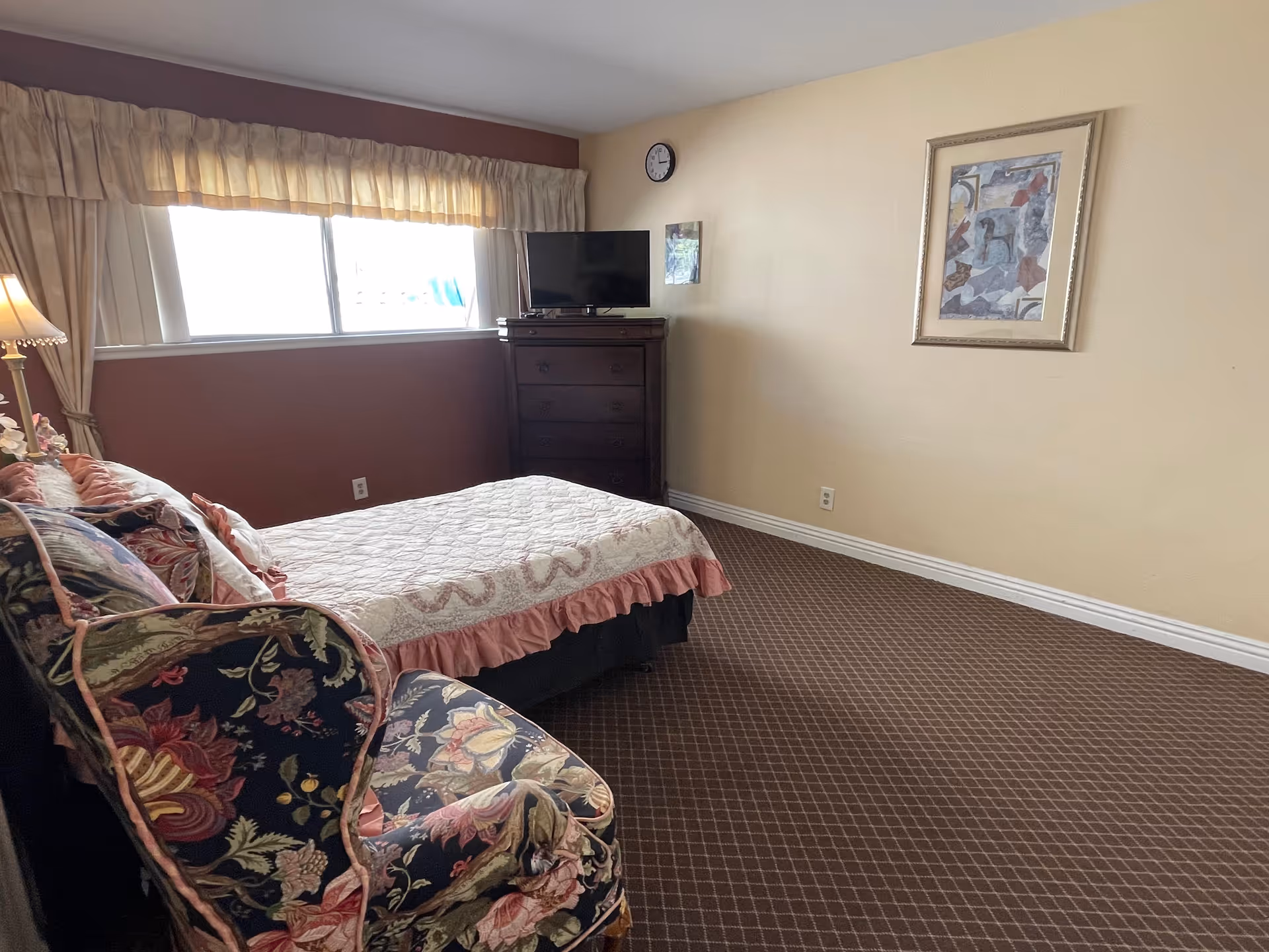 A bedroom in a senior living facility with a bed covered in a quilt, a floral upholstered armchair, a wooden dresser with a flat-screen TV on top, a window with curtains, a wall clock, and a framed abstract painting on the wall.