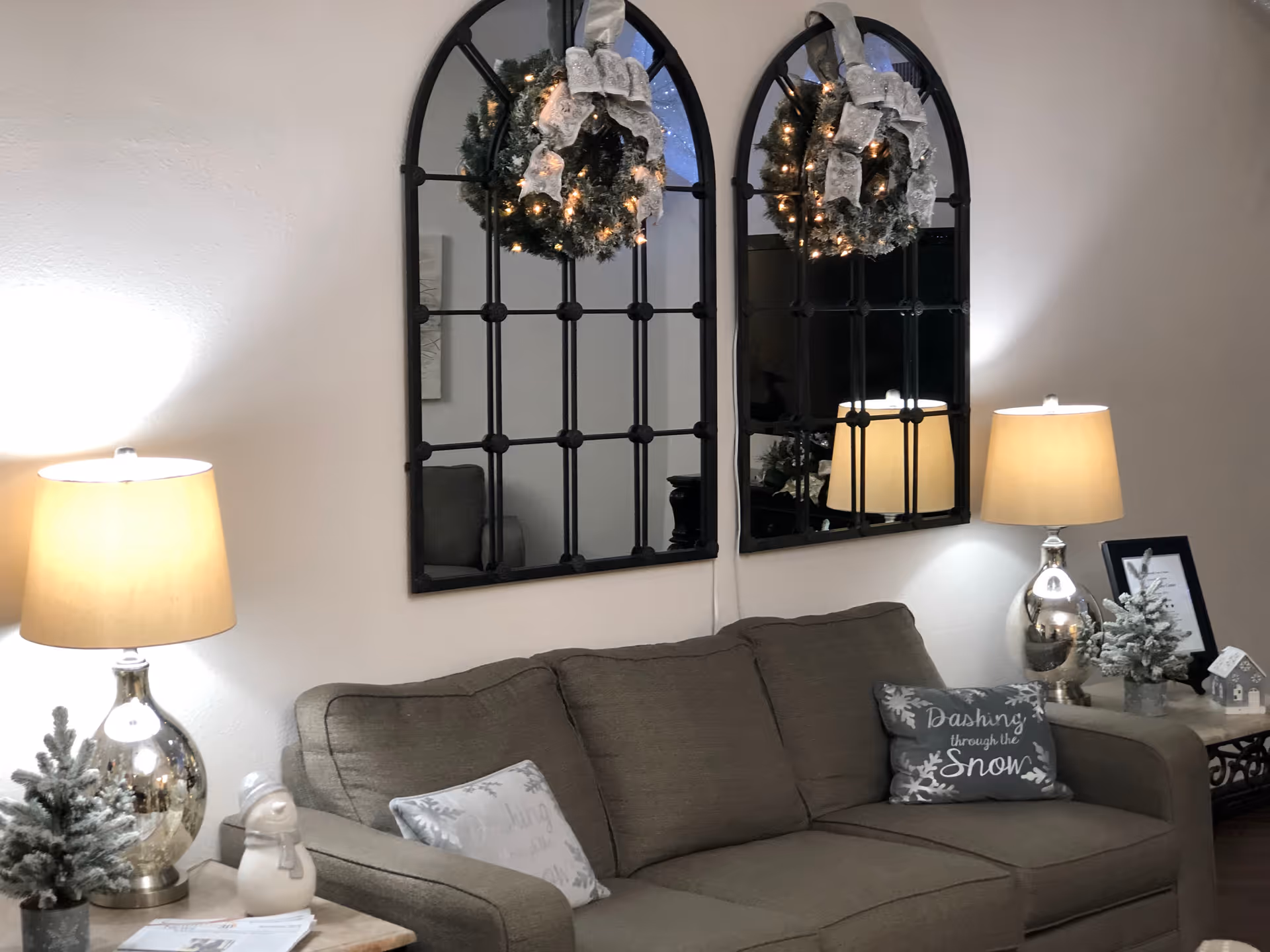 A cozy living room area with a gray sofa adorned with decorative pillows, including one that says 'Dashing through the Snow.' Two large arched mirrors with black frames hang on the wall above the sofa, each decorated with a festive wreath with lights and a silver bow. On either side of the sofa are side tables with silver lamps, small frosted Christmas trees, and holiday decorations including a small snowman figurine and a miniature house.