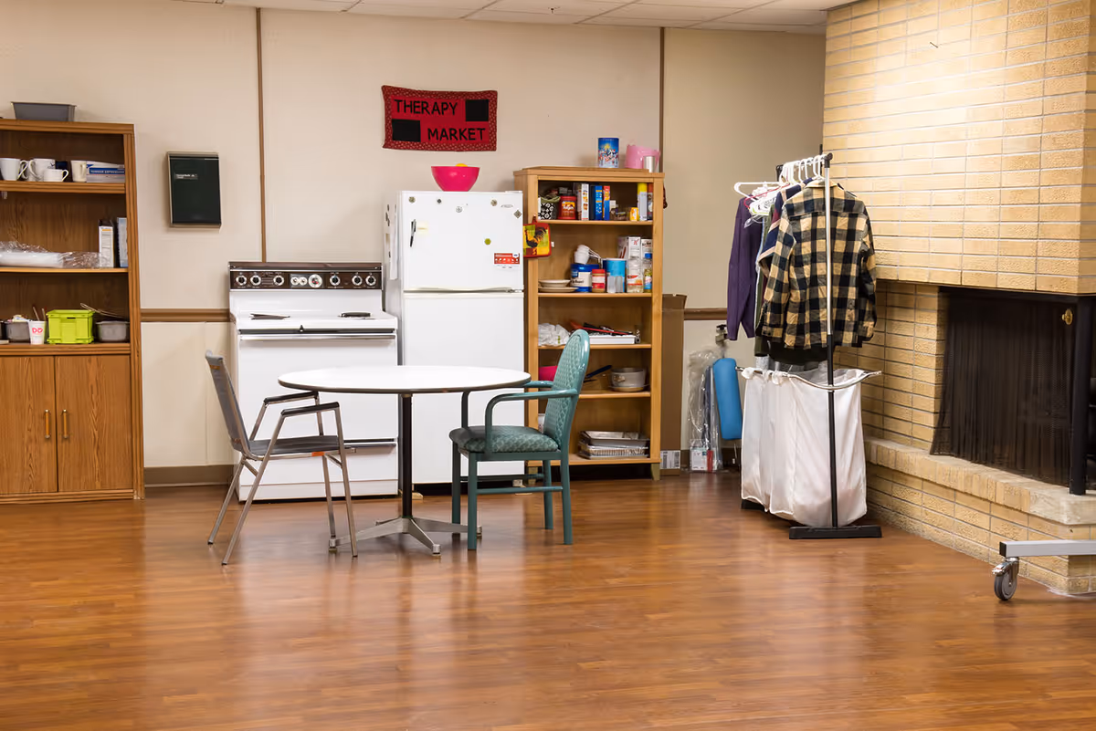 A room with a white round table and two chairs on a wooden floor. Behind the table is a white stove and refrigerator, with a wooden shelving unit filled with various food items. A red sign above the refrigerator reads 'THERAPY MARKET'. To the right, there is a clothing rack with jackets hanging and a brick fireplace.