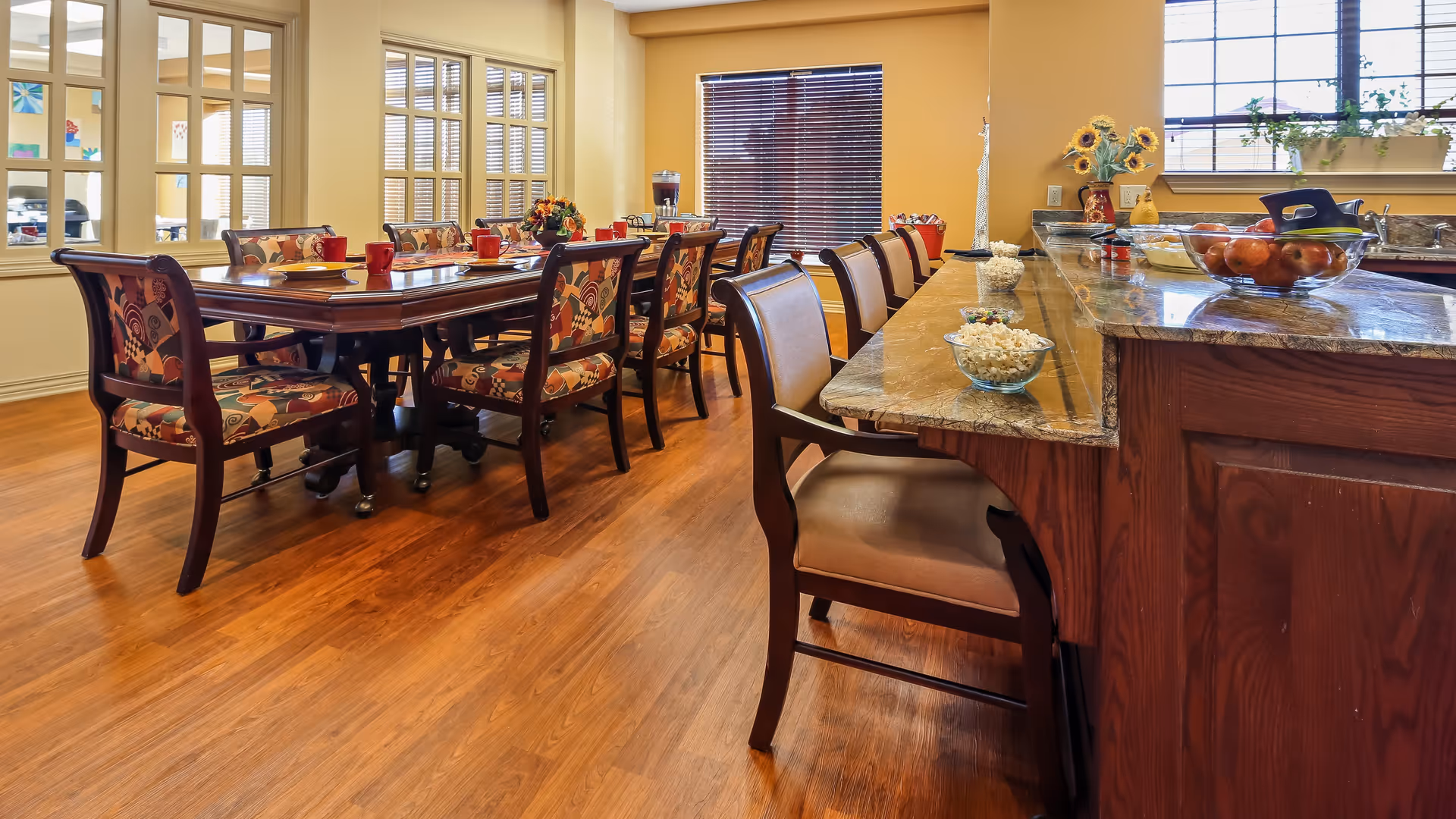 A warm and inviting dining area featuring a long wooden dining table with patterned cushioned chairs and a kitchen counter with high chairs. The counter has bowls of popcorn and a bowl of apples. Large windows with blinds allow natural light to fill the room, highlighting the wooden floor and yellow walls.