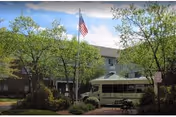 Exterior view of Meadows At Salem Run Senior Apartments showing a three-story building partially obscured by trees, an American flag on a flagpole, a senior shuttle bus parked in front, and a bench near the entrance.