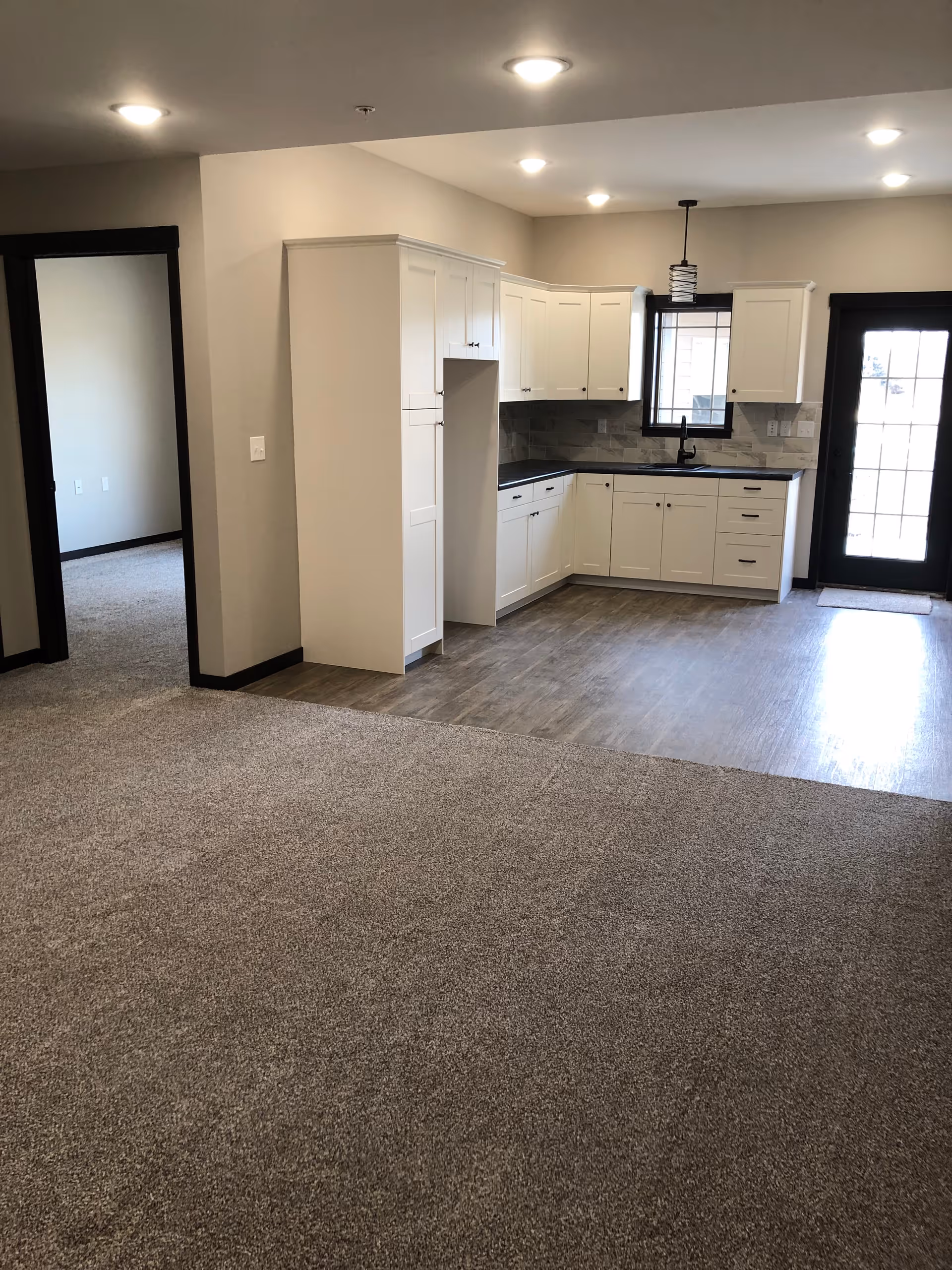 An interior view of a living space featuring a kitchen area with white cabinets, black countertops, a window above the sink, and a glass door leading outside. The floor transitions from carpet in the living area to wood-like flooring in the kitchen. There is a doorway leading to another room on the left.