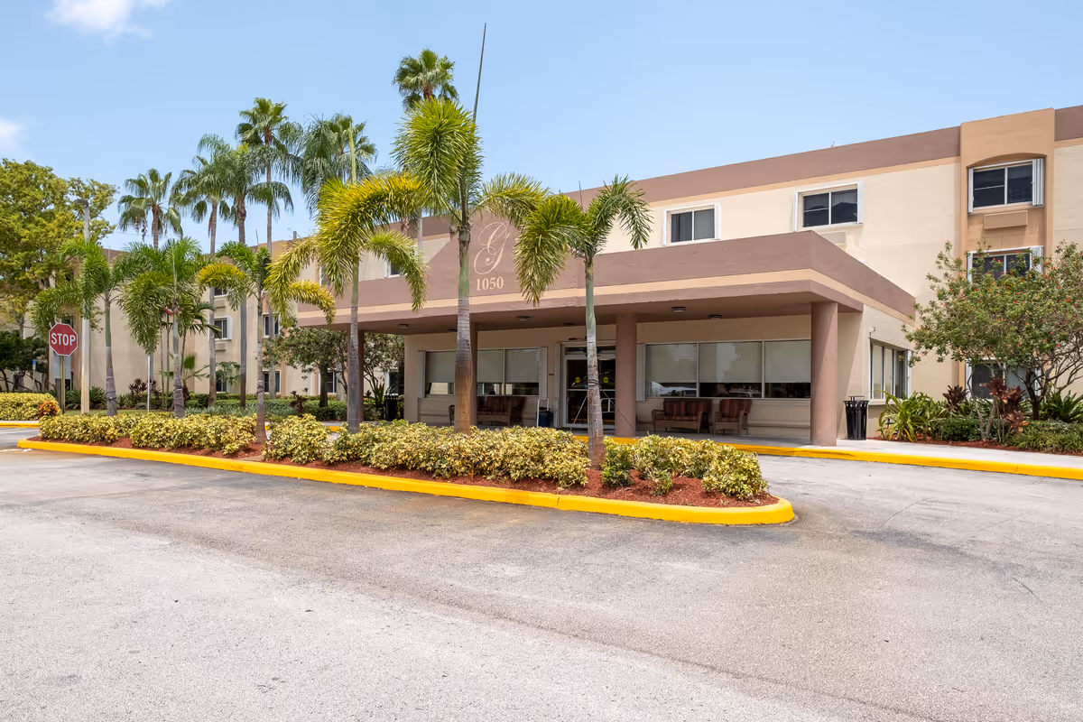 Exterior view of the entrance to a senior living facility named Grand Villa of Deerfield Beach, featuring a covered drop-off area, palm trees, shrubs, and a stop sign near the driveway.