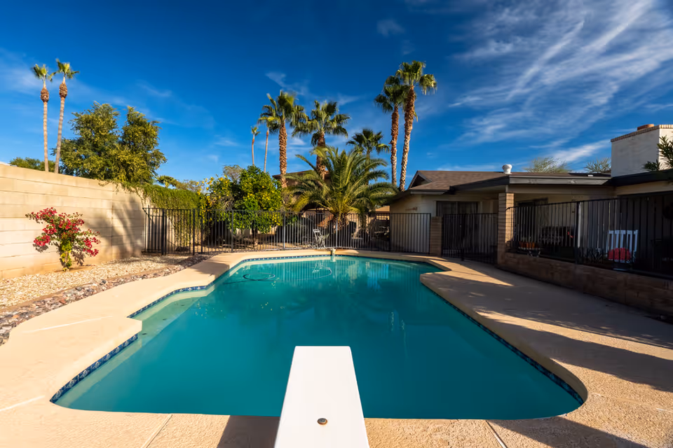 Outdoor swimming pool with a diving board in a fenced backyard area, surrounded by palm trees, bushes, and a clear blue sky. A single-story building with a covered patio is visible on the right side.