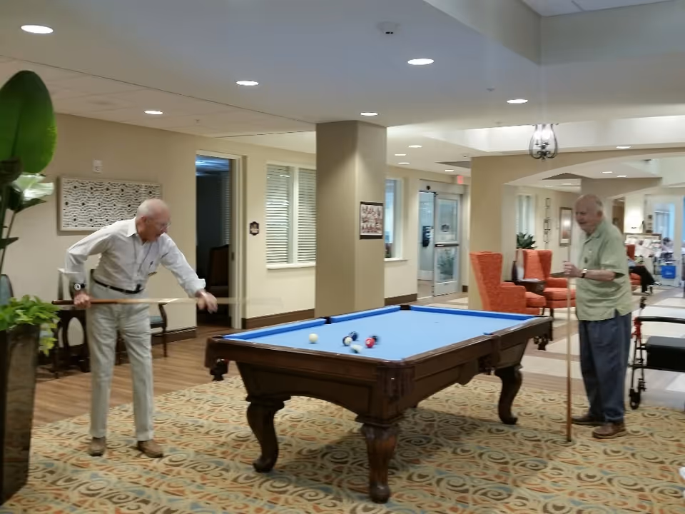 Two elderly men playing pool on a blue felt pool table in a well-lit common area with patterned carpet, beige walls, and orange armchairs in the background.