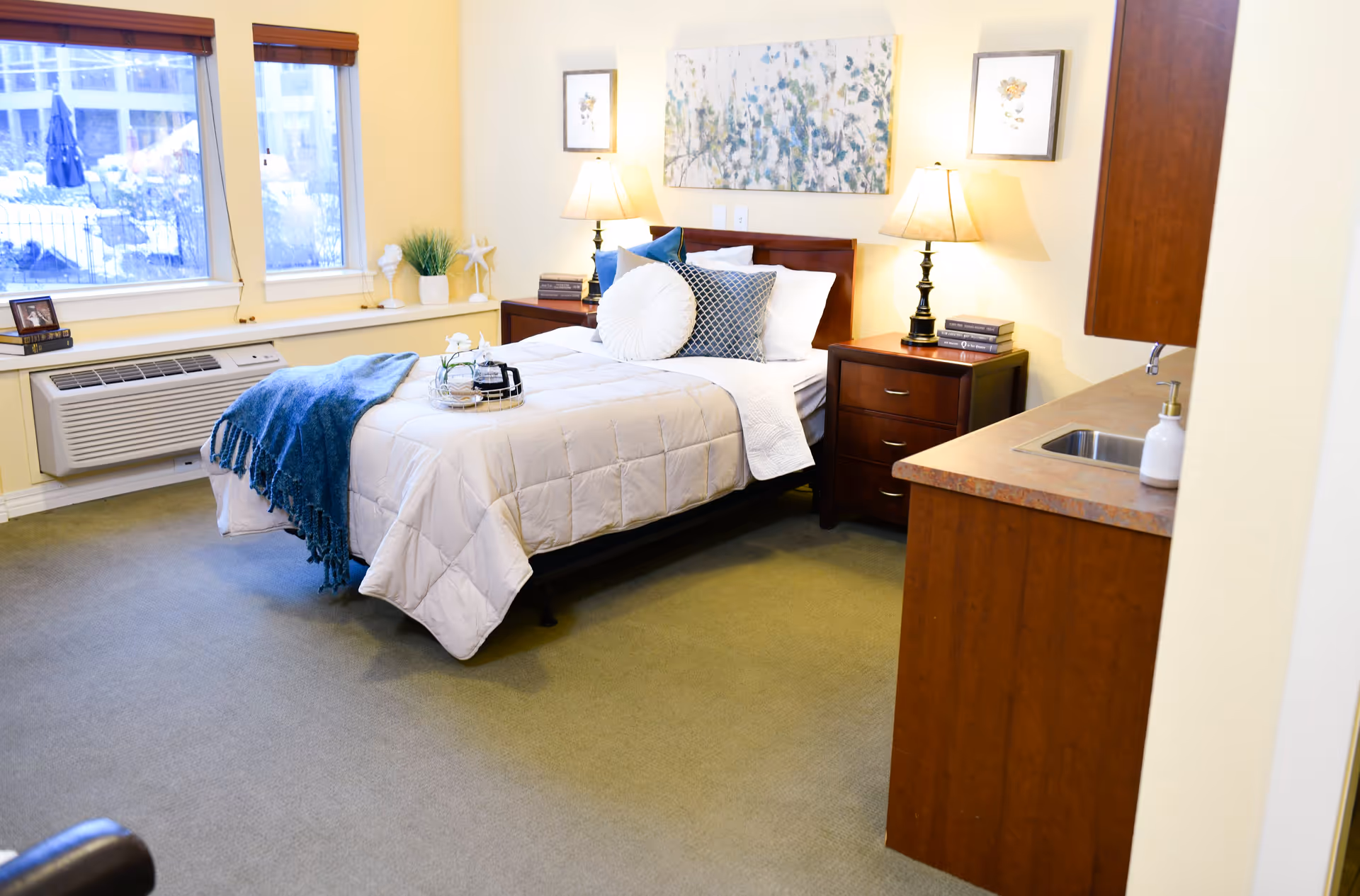 Well-lit furnished bedroom with a made bed, nightstands and lamps, windows on the left and a small kitchenette area on the right.