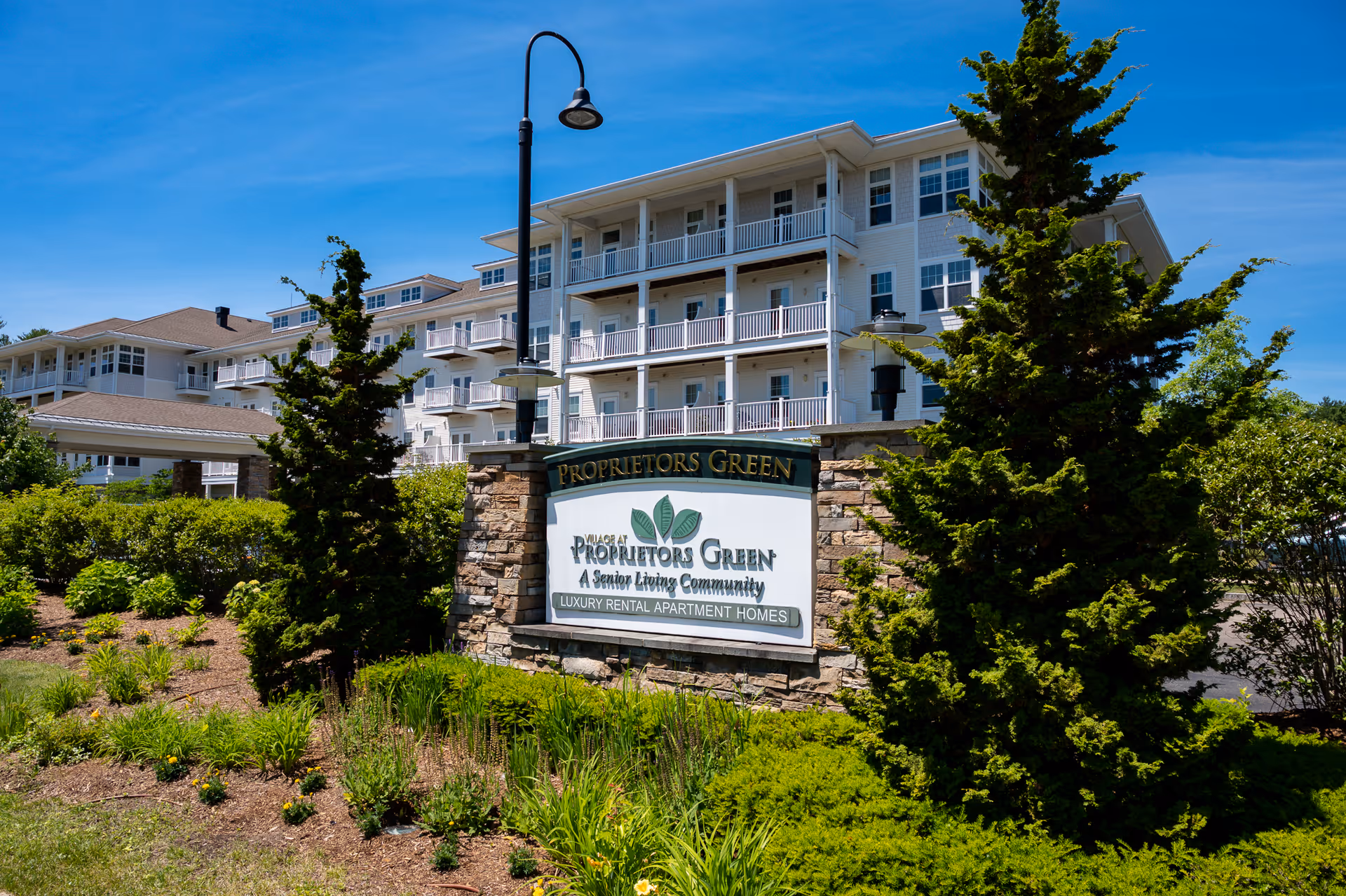 Exterior view of the Village At Proprietors Green senior living community building with a large sign in front surrounded by greenery and trees under a clear blue sky.