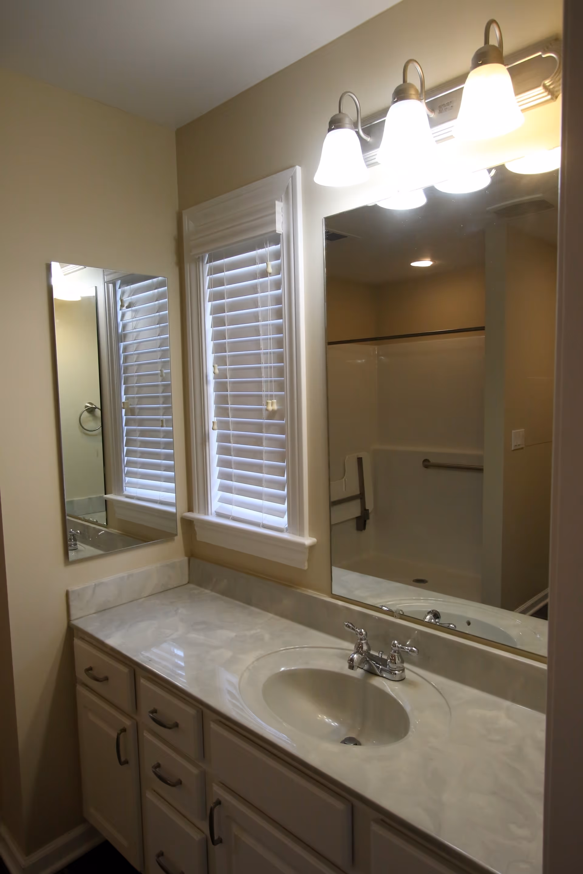 Bathroom vanity with a marble countertop, an oval sink with chrome faucet, a large mirror above the sink, and a window with white blinds. The reflection in the mirror shows a shower area with a grab bar and a fold-down seat. The bathroom walls are painted beige.