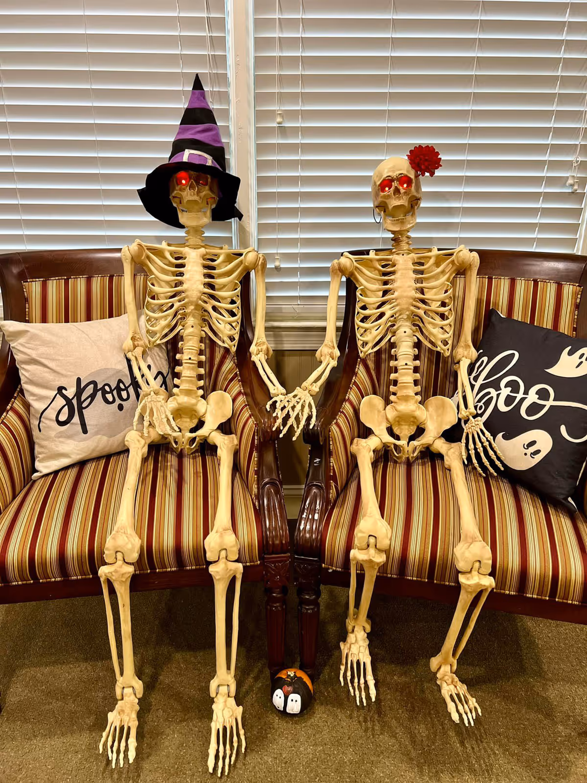 Two seated Halloween skeleton decorations with glowing red eyes and hats holding hands on striped chairs in a room with blinds and festive pillows.