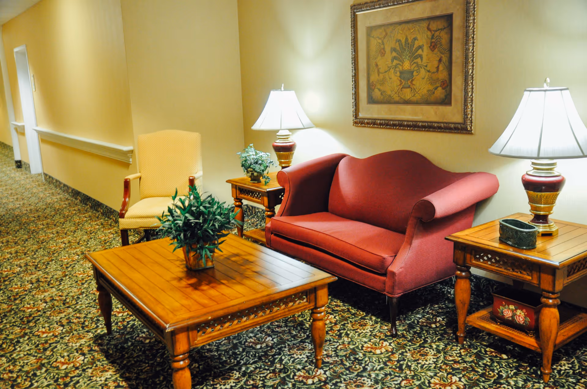 A cozy sitting area in a hallway with a red upholstered loveseat, a yellow armchair, two wooden side tables with lamps, a wooden coffee table with a potted plant, and a framed artwork on the wall. The floor is covered with a patterned carpet.