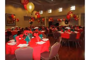 A decorated dining room with round tables covered in red tablecloths, each set with plates, napkins folded in a festive manner, and silverware. Red and gold balloons are tied to the chairs and floating above the tables. The room has several windows near the ceiling and framed artwork on the walls.