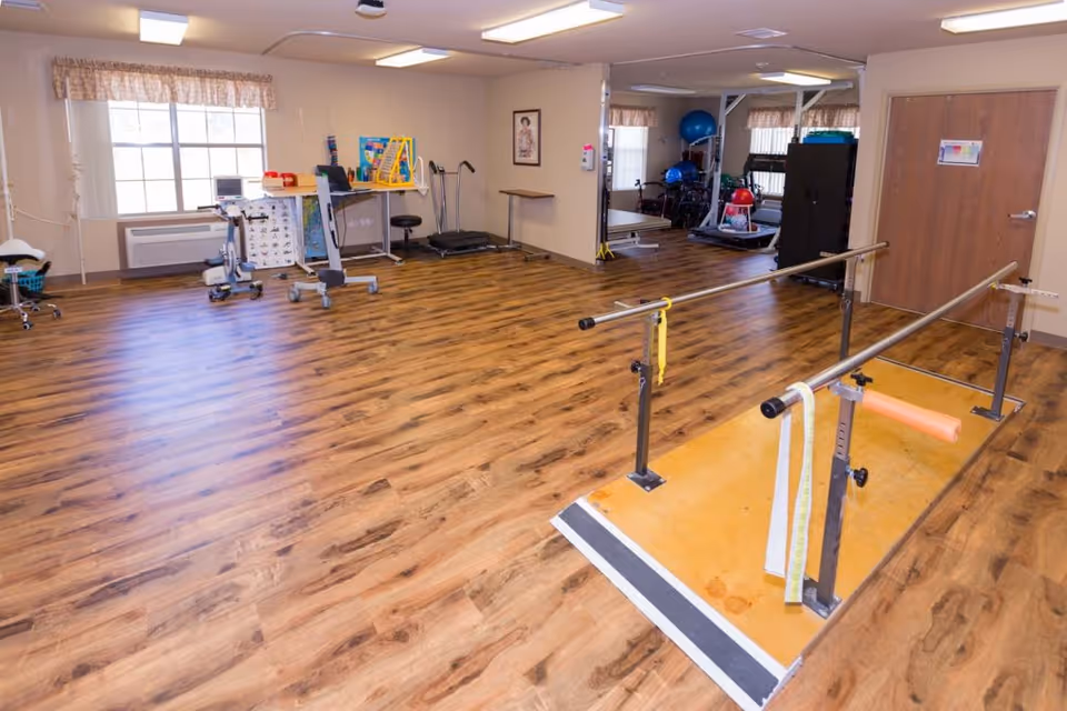 A physical therapy room with parallel walking bars, exercise equipment, and wood-look flooring.