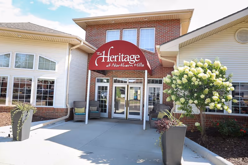 Entrance of The Heritage at Northern Hills facility with a red awning displaying the facility name above double glass doors. There are two wicker chairs with cushions on either side of the entrance, large planters with greenery, and a blooming white flower tree to the right. The building exterior is a combination of brick and light-colored siding with multiple windows.