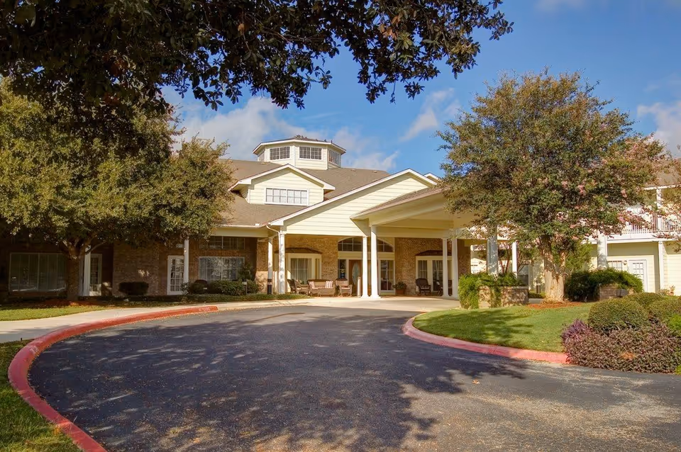Exterior view of The Waterford on Huebner senior living facility showing the front entrance with a covered driveway, surrounded by trees and landscaped greenery under a blue sky with some clouds.