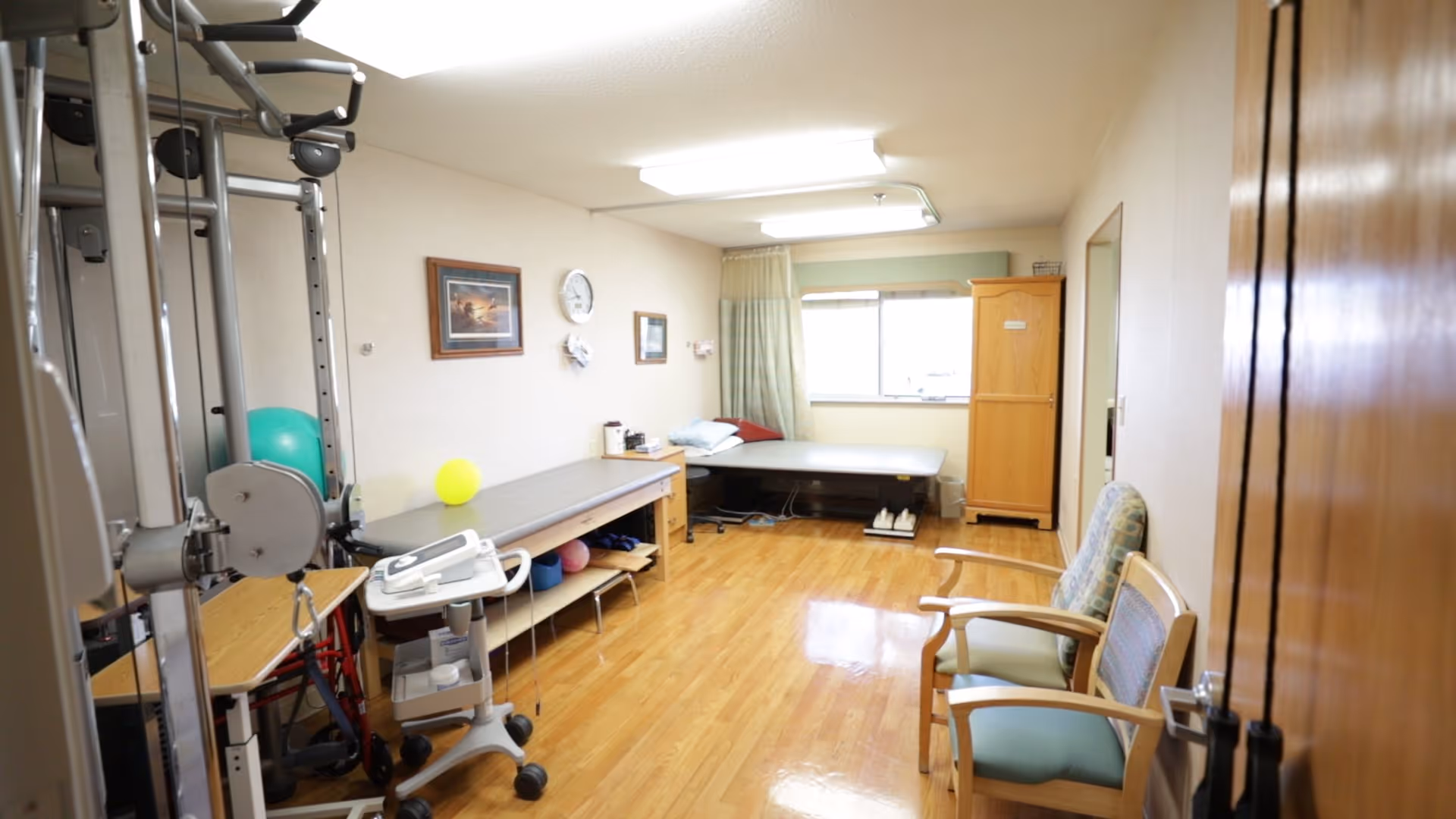 A therapy or rehabilitation room with exercise equipment, a treatment table, chairs, and a wooden cabinet. The room has wooden flooring, a window with curtains, and framed pictures on the wall.