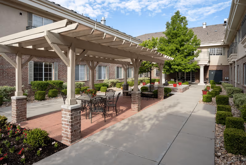 Outdoor courtyard area at Escalante At Coventry featuring a covered pergola with metal chairs and tables, surrounded by neatly trimmed bushes, potted plants, and a tree, with the building facade in the background under a partly cloudy sky.