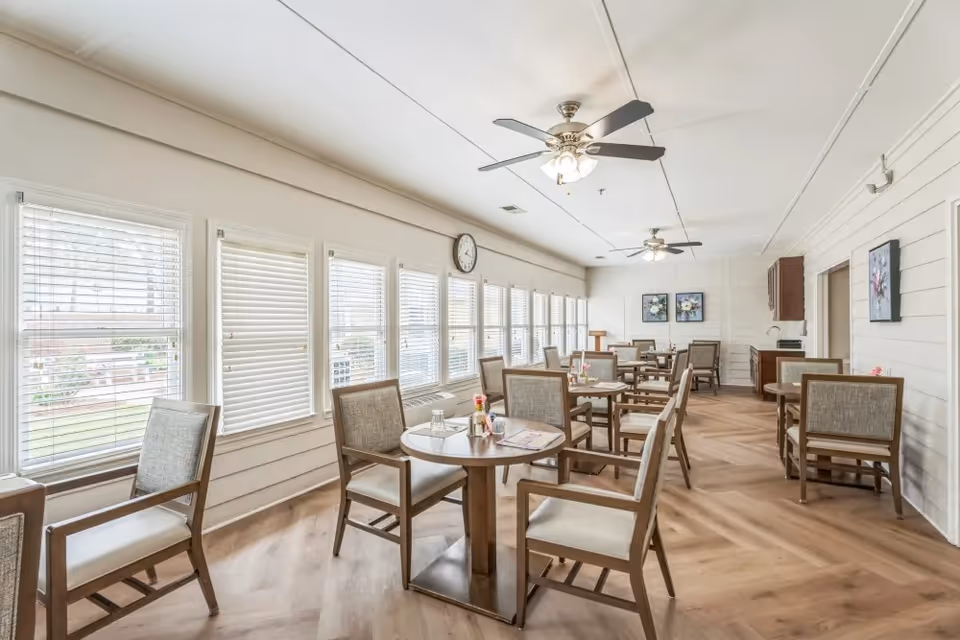 A bright dining room with multiple round tables and cushioned chairs arranged neatly. Large windows with white blinds line one wall, allowing natural light to fill the space. The room has light-colored wooden flooring, ceiling fans with lights, and floral artwork on the walls.