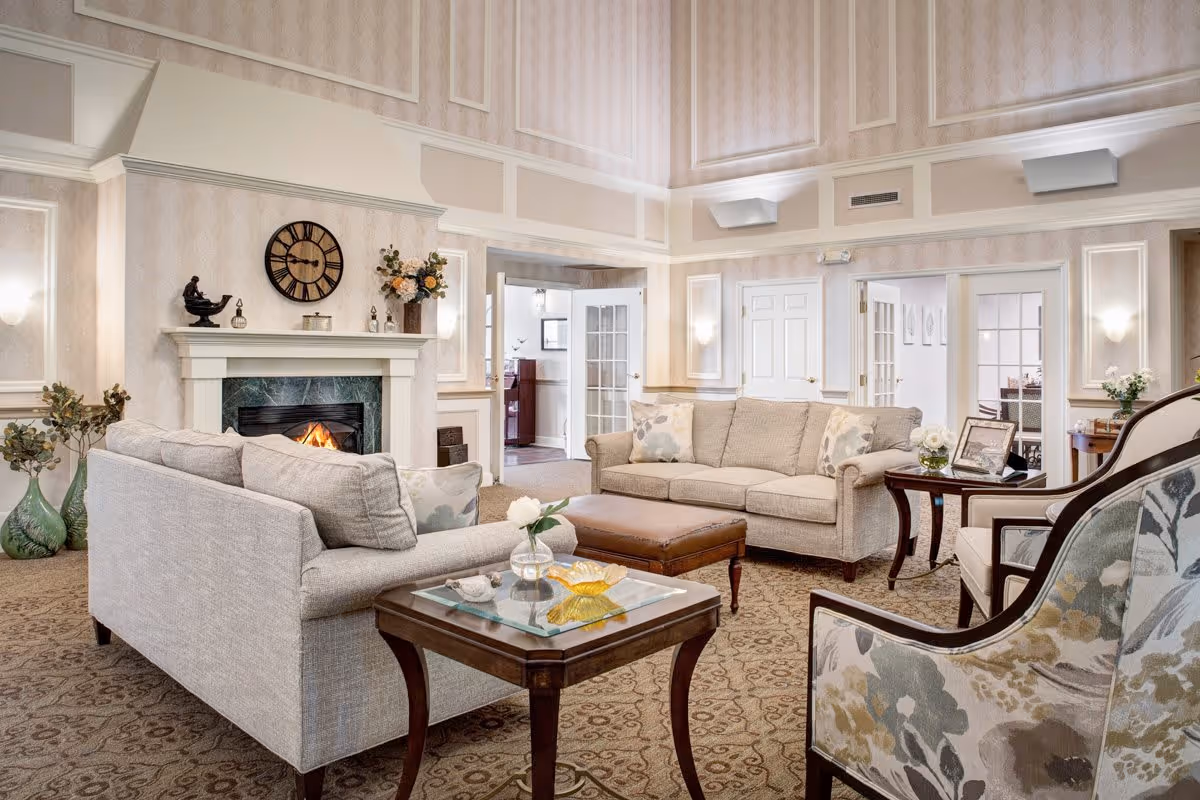 A spacious and elegantly decorated living room with two beige sofas, a floral upholstered armchair, a wooden coffee table with glass top, and a brown leather ottoman. The room features a lit fireplace with a clock and decorative items on the mantel, patterned carpet, and soft wall lighting. There are doorways leading to other rooms in the background.