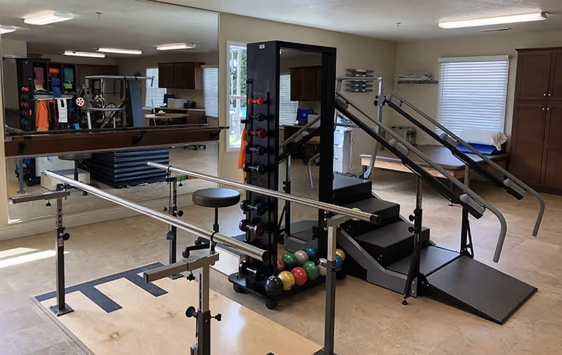 A rehabilitation exercise room with parallel bars for walking practice, a rack of colorful dumbbells, a set of stairs with handrails for physical therapy, a large mirror on one wall, and windows letting in natural light.