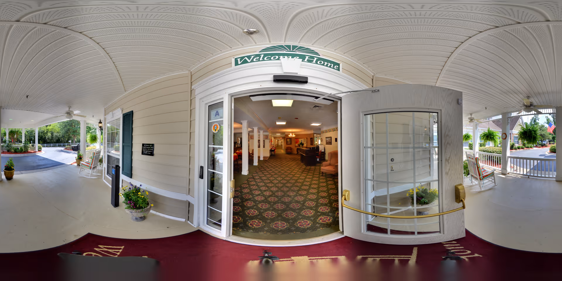 Covered entrance porch with rocking chairs and open double doors beneath a 'Welcome Home' sign revealing the facility lobby.