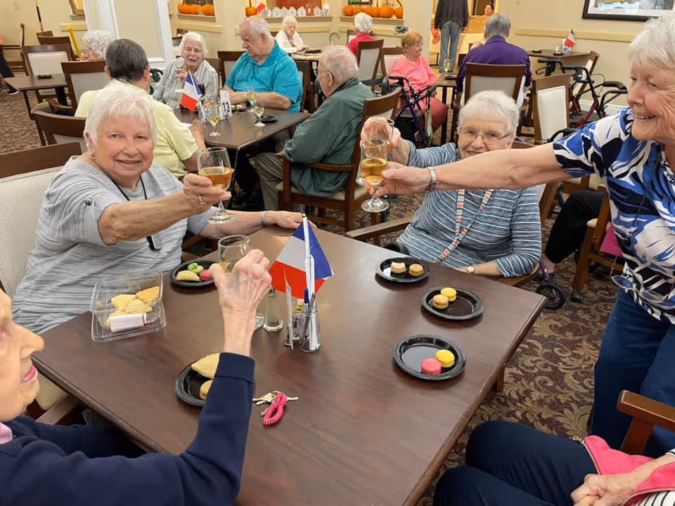 A group of elderly people sitting around tables in a communal dining area, smiling and raising glasses in a toast. Plates with cookies and macarons are on the tables, and small flags are placed in the center. The room is decorated with pumpkins and has a warm, welcoming atmosphere.