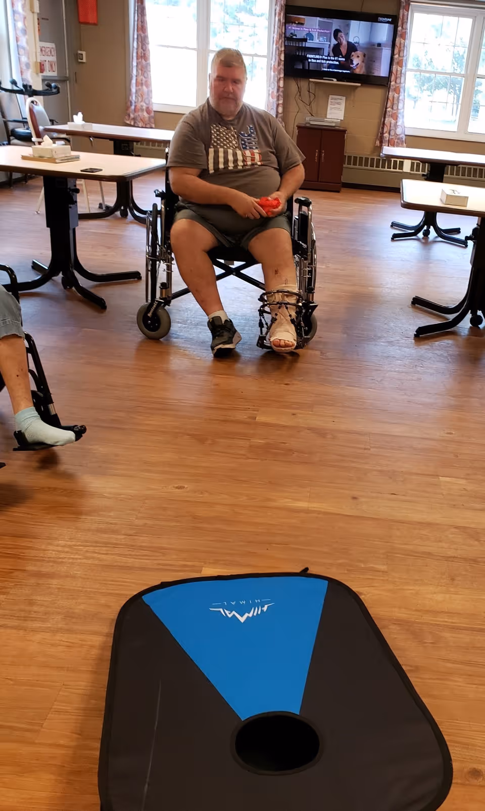 A man in a wheelchair with a leg cast and brace is sitting in a room with wooden flooring, holding a red bean bag. There are tables and chairs around, a television mounted on the wall, and windows with curtains letting in natural light.