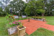 Outdoor patio area with red brick paving, two metal swing benches, a birdbath, and raised garden beds with flowers and greenery, surrounded by a grassy lawn and large trees in the background.