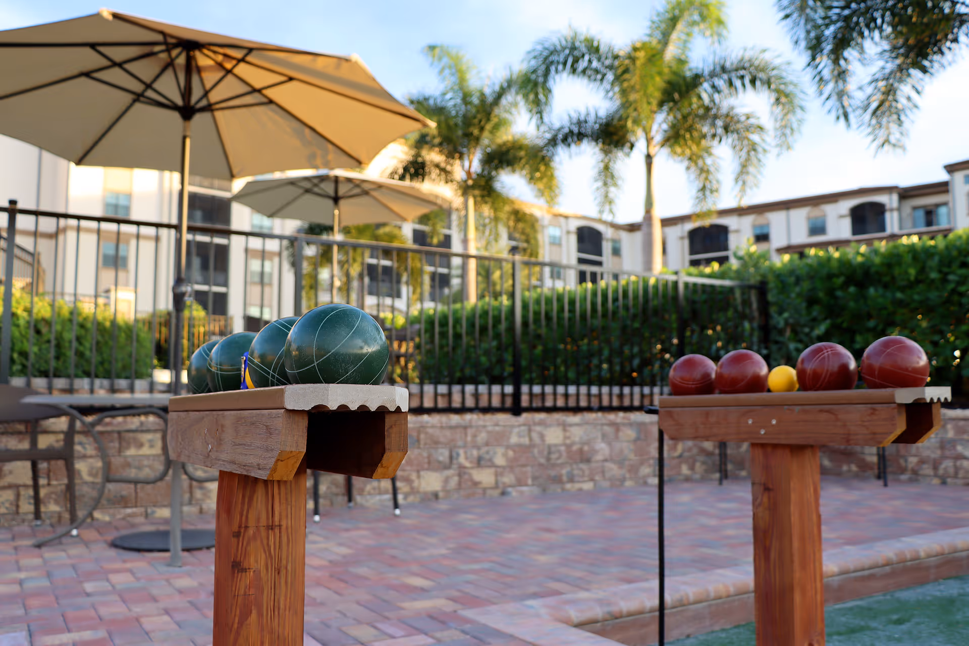 Outdoor bocce ball court with bocce balls arranged on wooden racks, patio umbrellas, palm trees, and a multi-story building in the background.