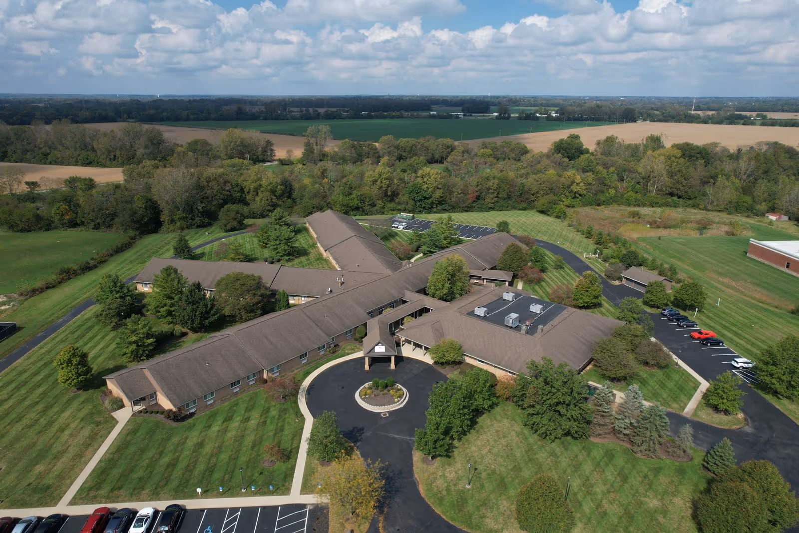Aerial view of a single-story senior living facility named Our Home - Fairborn, surrounded by well-maintained lawns, trees, and parking areas, with fields and forested land in the background under a partly cloudy sky.