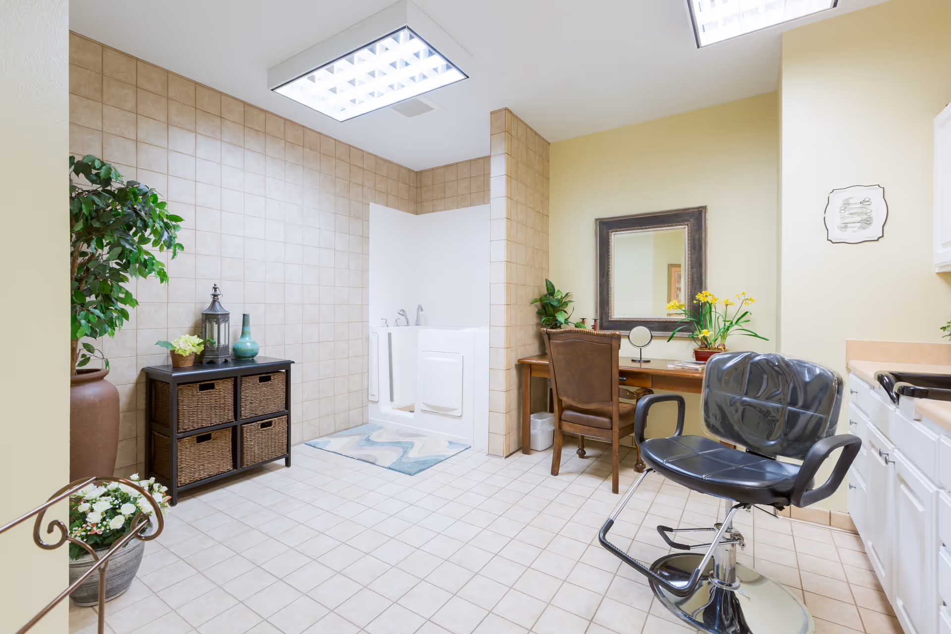 A spacious, well-lit room with beige tiled floors and walls. The room features a walk-in bathtub with a door, a black salon chair, a wooden desk with a mirror and plants, a small cabinet with wicker baskets, and decorative plants placed around the room.
