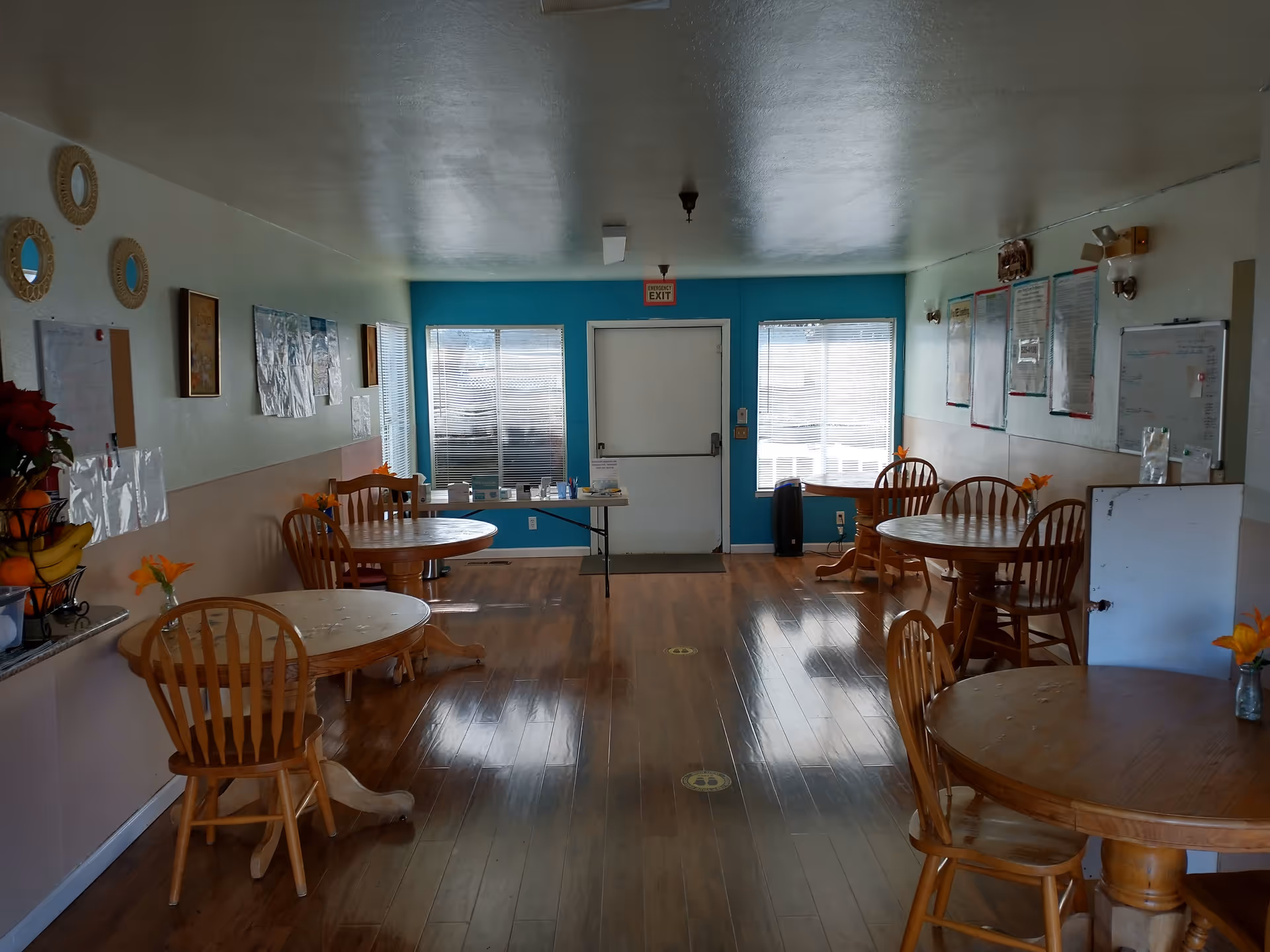 Interior view of a dining room with wooden round tables and chairs arranged along the walls. The room has wooden flooring, light-colored walls with some decorations and notices, and a door with windows on either side at the far end. There are small vases with orange flowers on the tables.