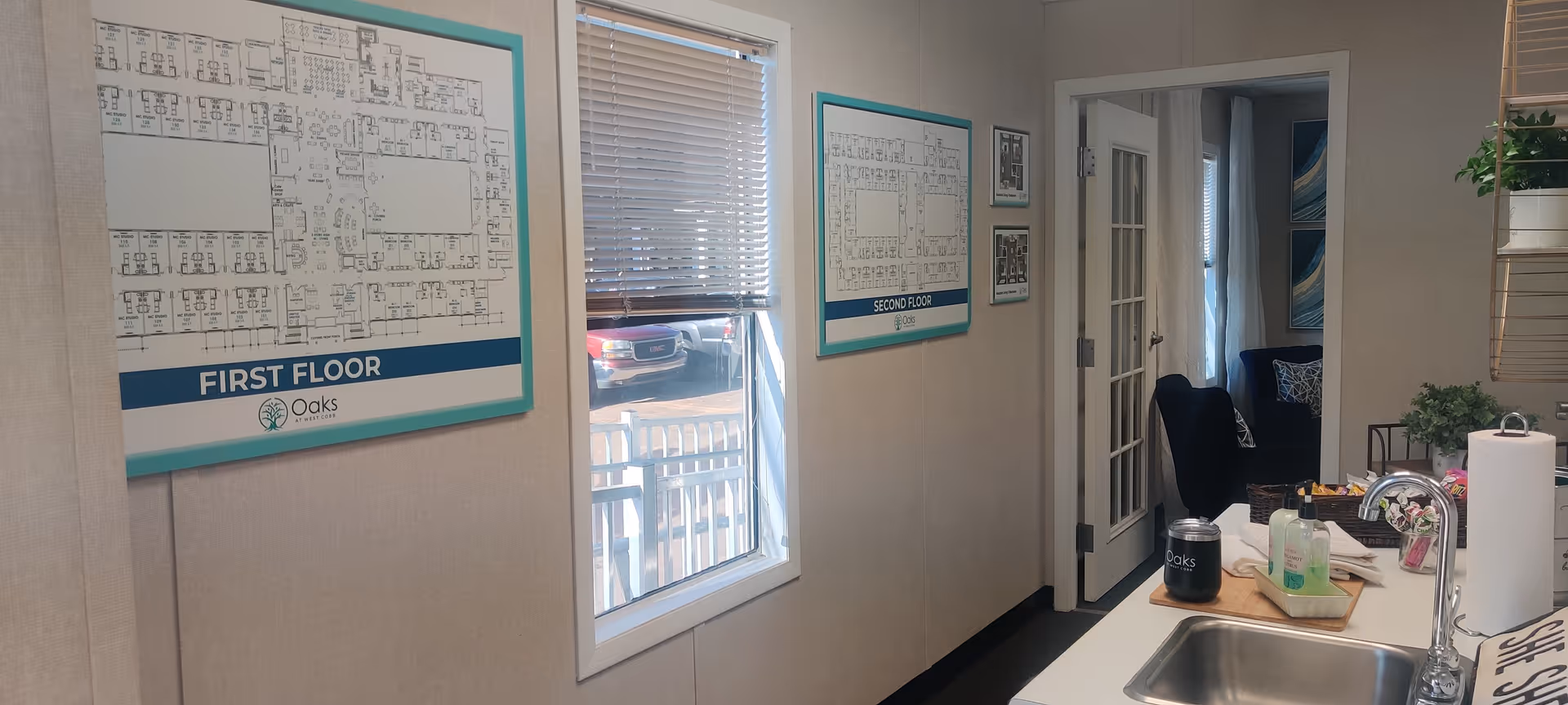 Interior view of a room in Oaks at West Cobb facility showing two framed floor plans labeled First Floor and Second Floor on the wall, a window with blinds partially open, a doorway leading to a room with chairs and artwork, and a kitchen counter with a sink, soap dispenser, paper towel roll, and a cup with the Oaks logo.