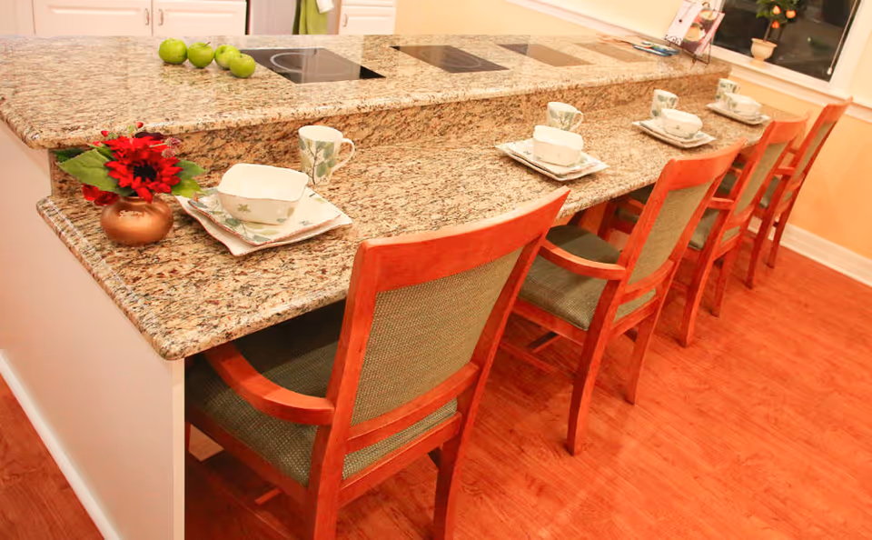 A kitchen counter with a granite countertop featuring four wooden chairs with green cushions lined up along the counter. The counter is set with plates, bowls, cups, and saucers, and there is a small vase with red flowers and a group of green apples on the countertop. The floor is wooden, and there is a window in the background.