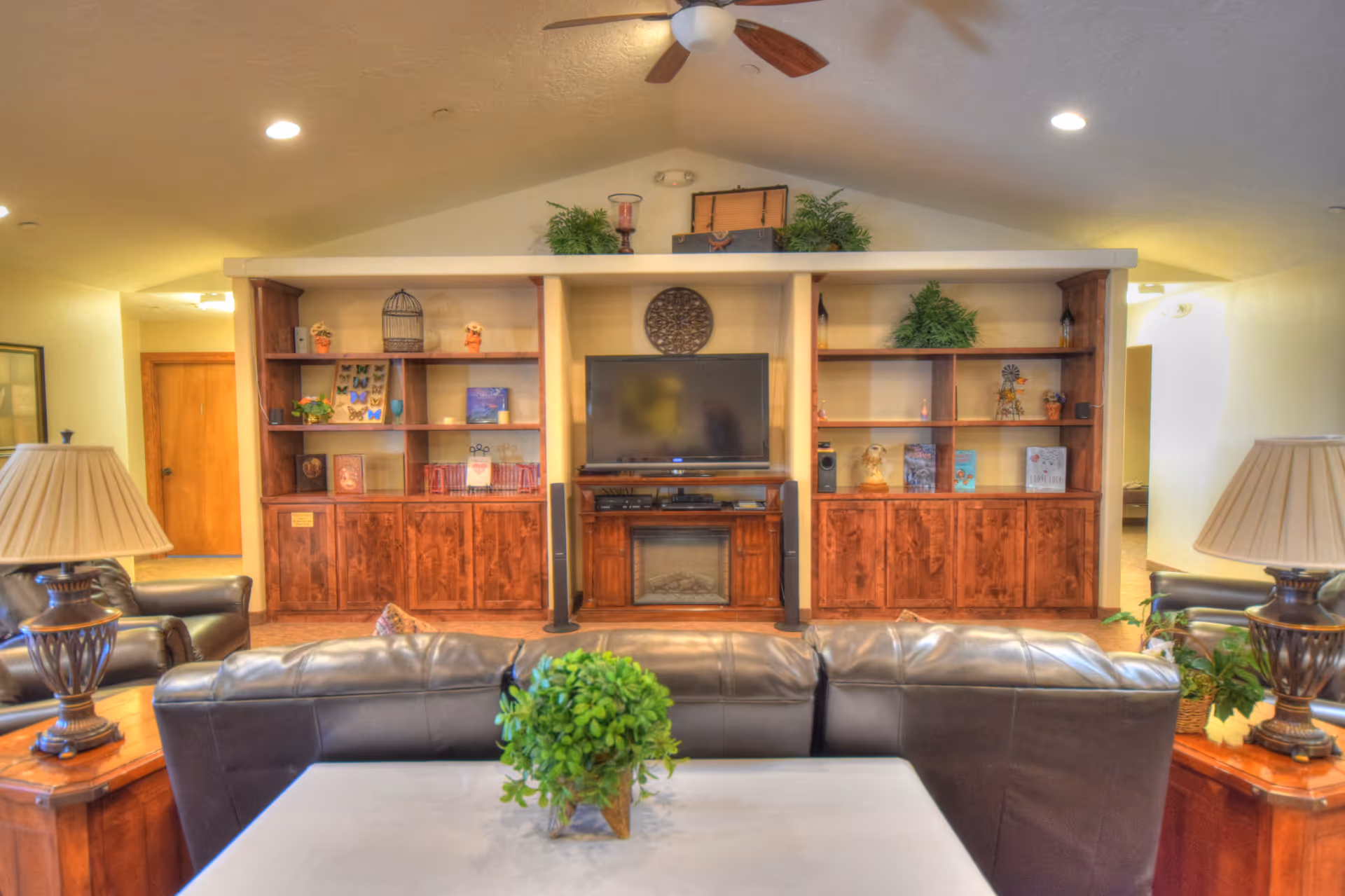 A cozy living room with a leather sofa and armchairs facing a wooden entertainment center with a flat-screen TV and electric fireplace. The shelves on either side of the TV are decorated with plants, books, and decorative items. Two table lamps with beige shades are placed on wooden side tables beside the sofa. A ceiling fan with wooden blades is mounted on the ceiling.
